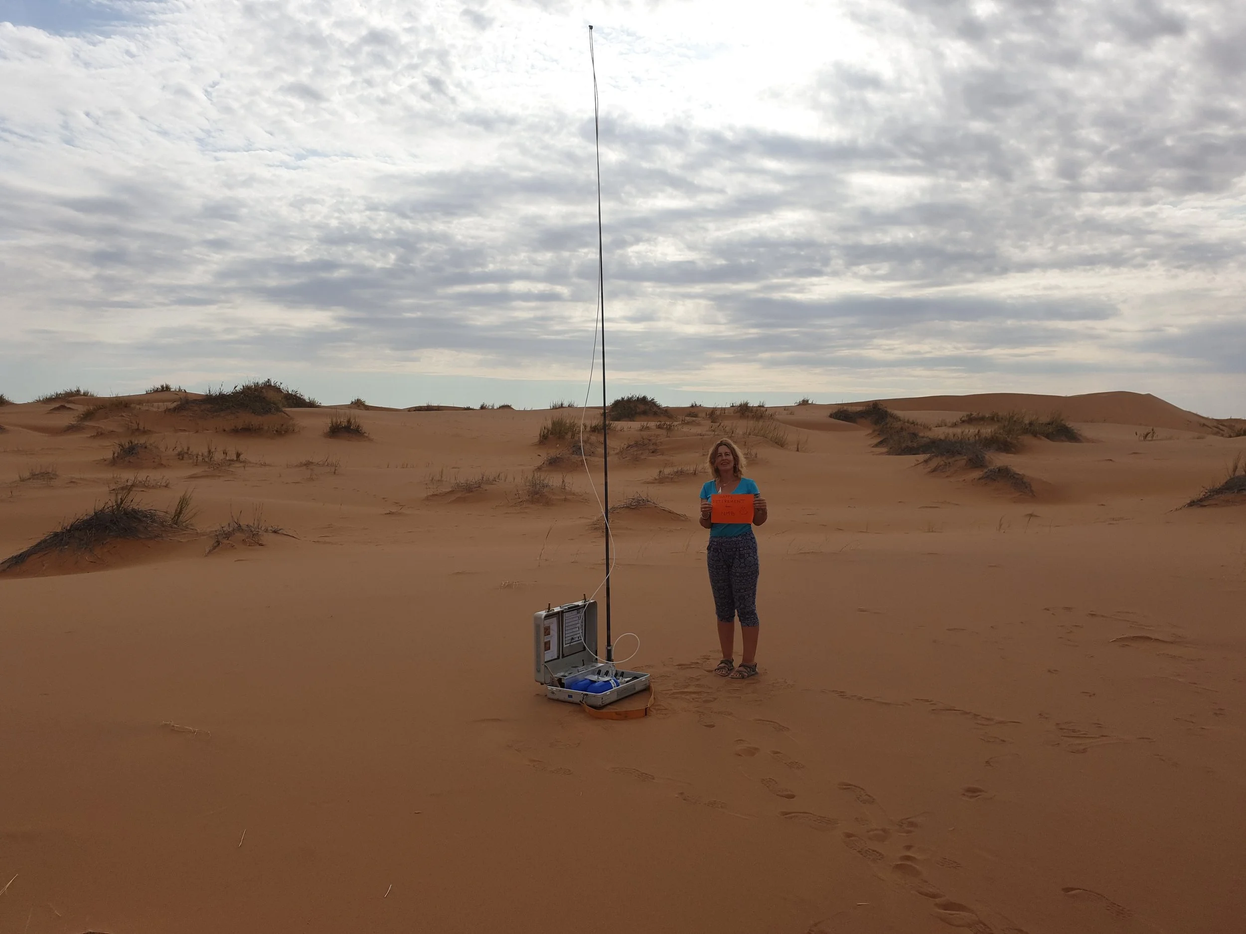 A woman in a blue shirt and patterned pants stands in a desert landscape holding an orange sign, with equipment on the ground nearby connected to a tall antenna, under a cloudy sky.