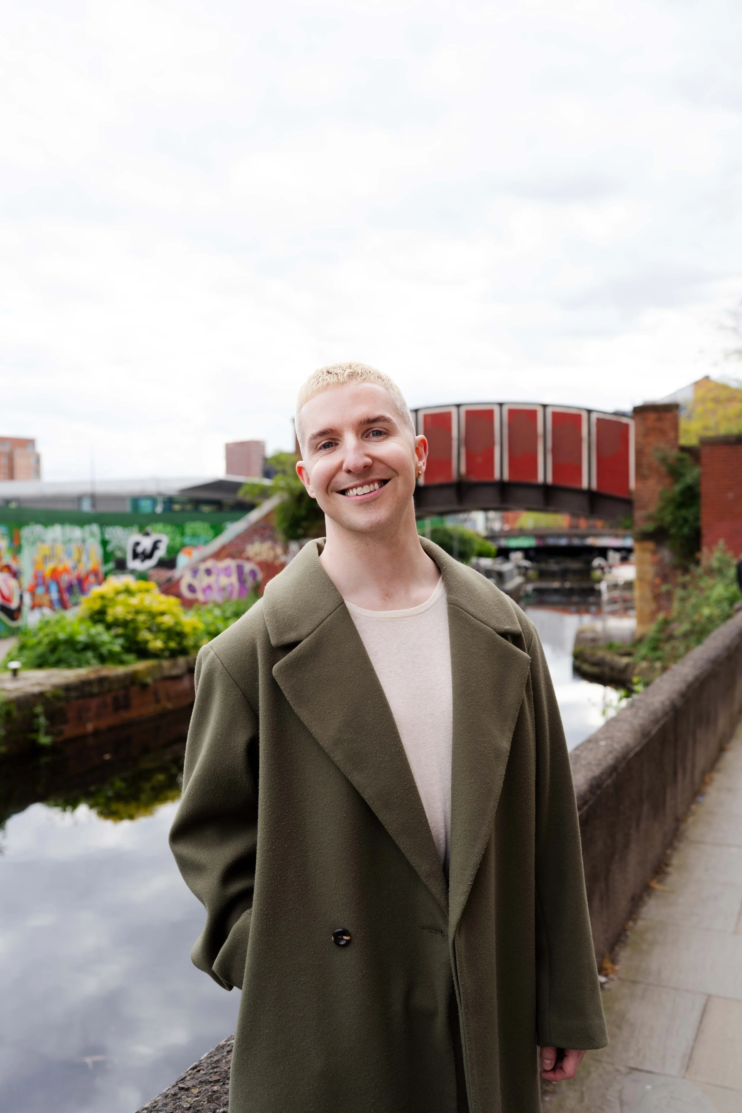 A young person with short blonde hair, wearing a green coat, standing outdoors near a canal with graffiti-covered walls, a small bridge, and buildings in the background during daytime with cloudy sky.