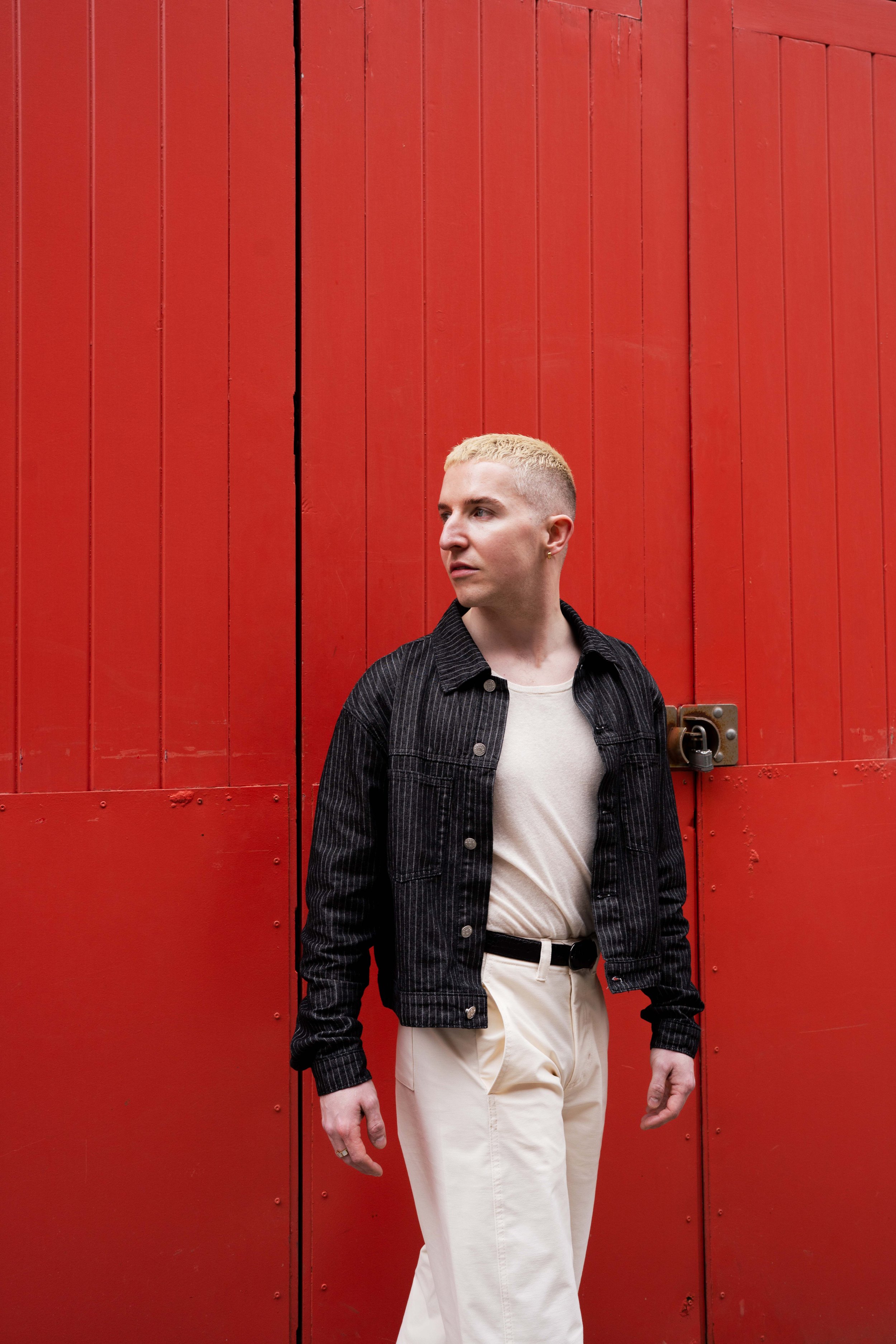 A person with short blonde hair wearing a black jacket over a cream shirt, standing in front of a large red wooden door.