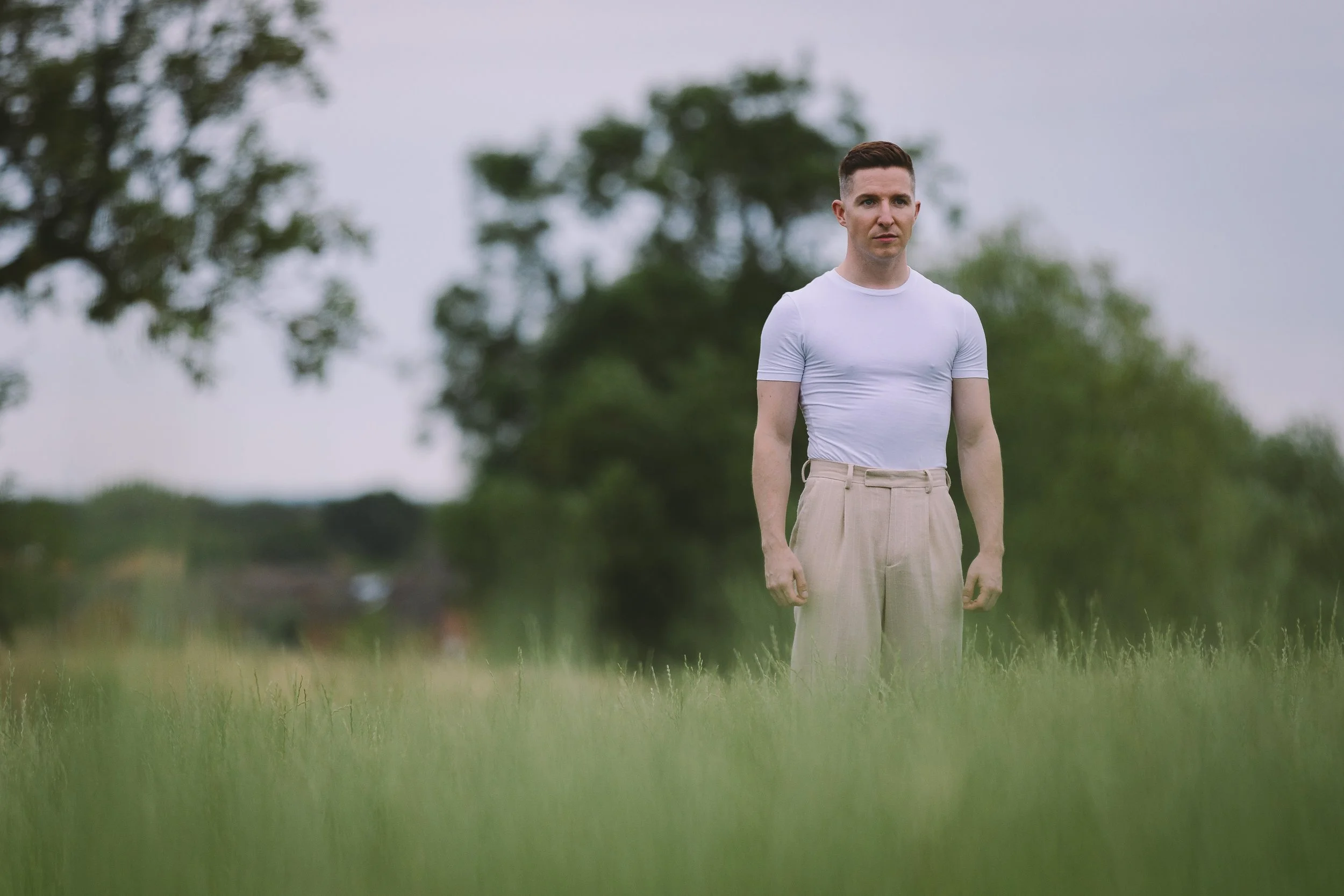 A man standing in a grassy field with trees in the background, wearing a white t-shirt and beige pants.