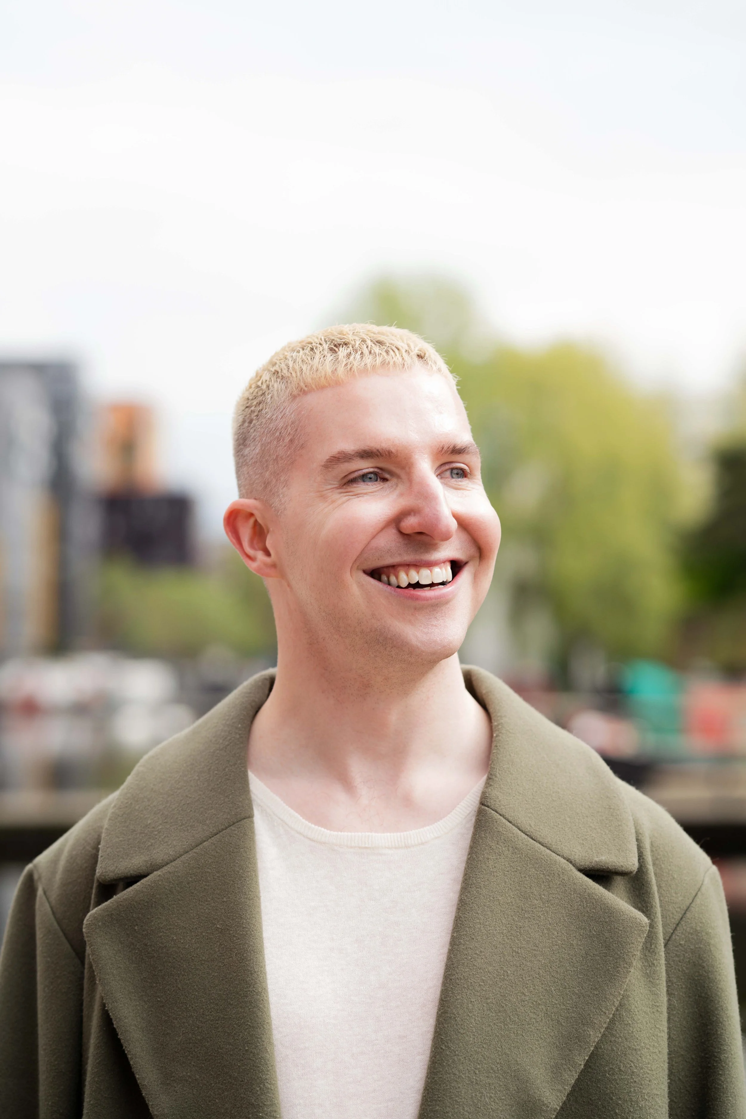 Smiling young man with short blond hair wearing a beige coat and white shirt outdoors.