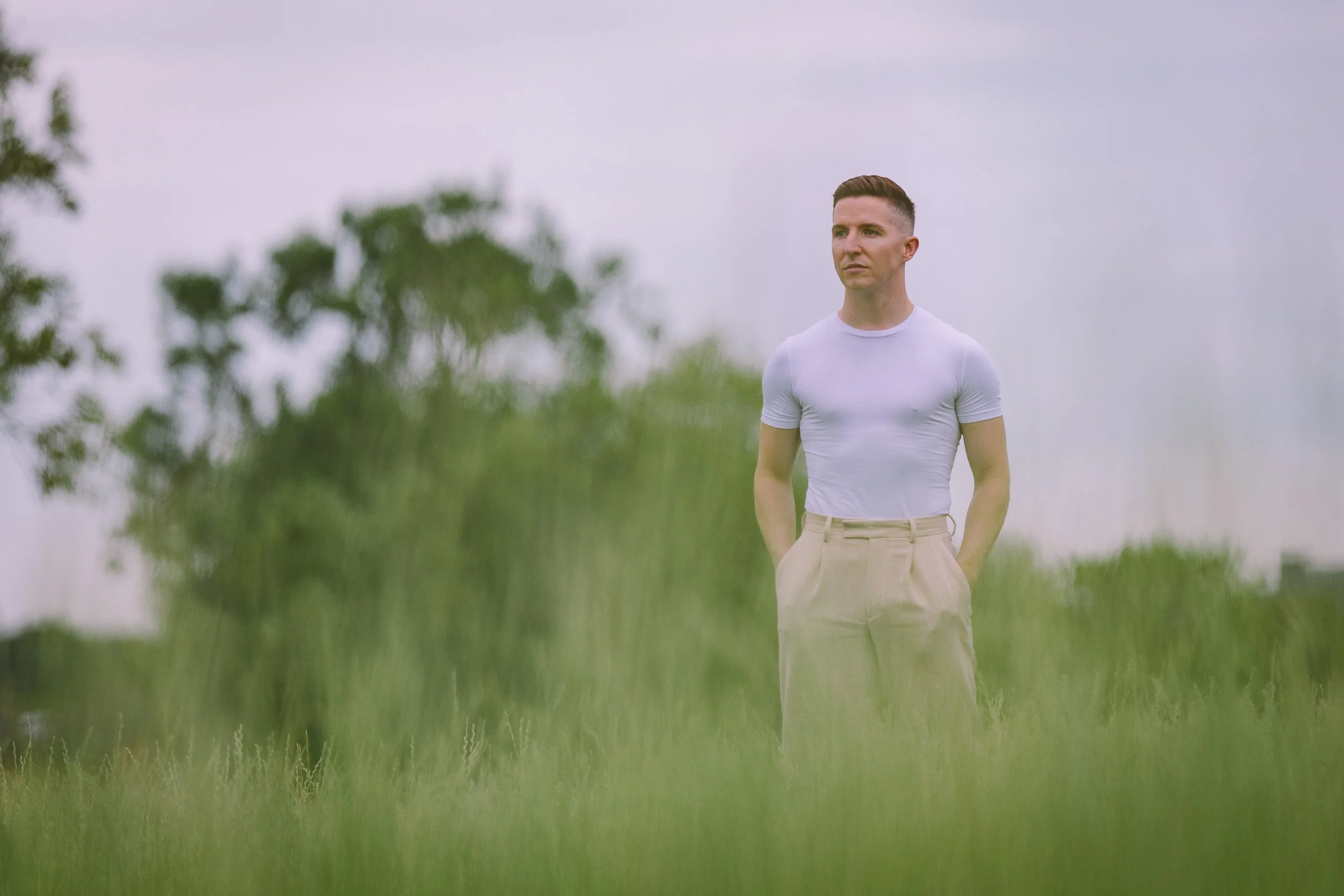 A young man in a white t-shirt and beige pants stands outdoors in a grassy area, looking into the distance with a thoughtful expression.