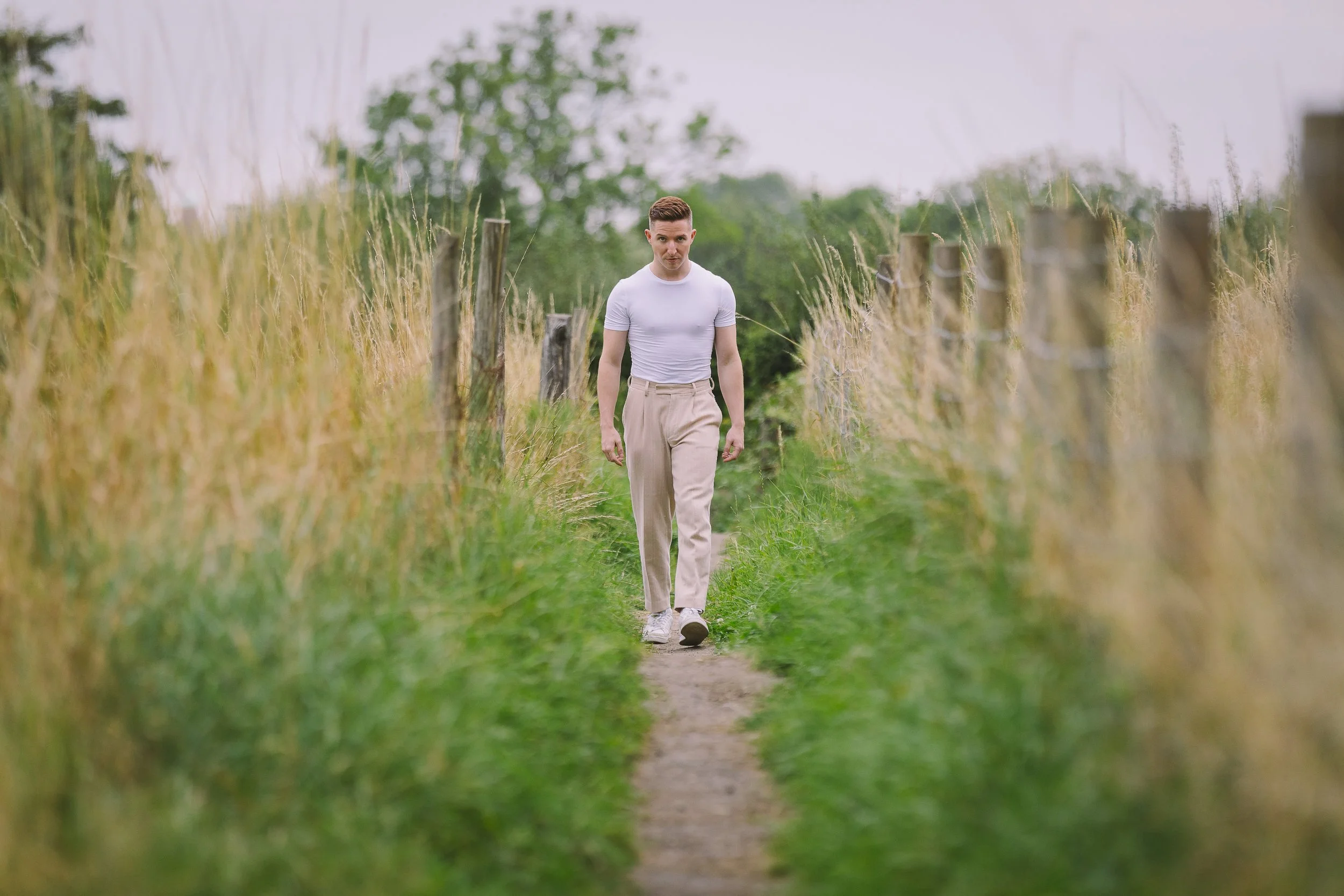 A young man walking down a narrow dirt path between two fences in a lush green outdoor setting, wearing a white t-shirt and beige pants.
