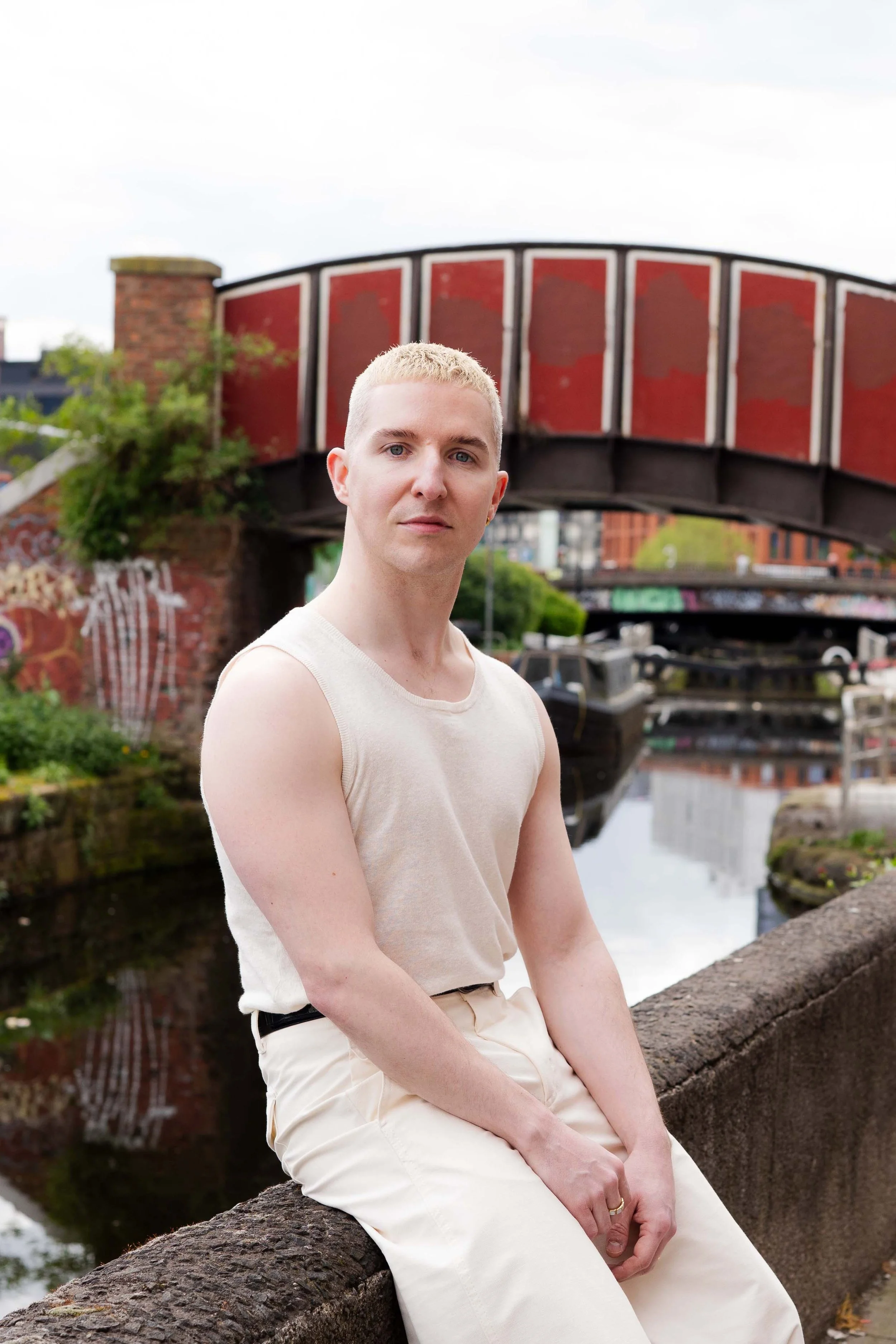 A person with short blond hair, wearing a sleeveless beige top and cream-colored pants, sitting on a stone ledge near a canal with boats, an arched red brick bridge, and city buildings in the background.