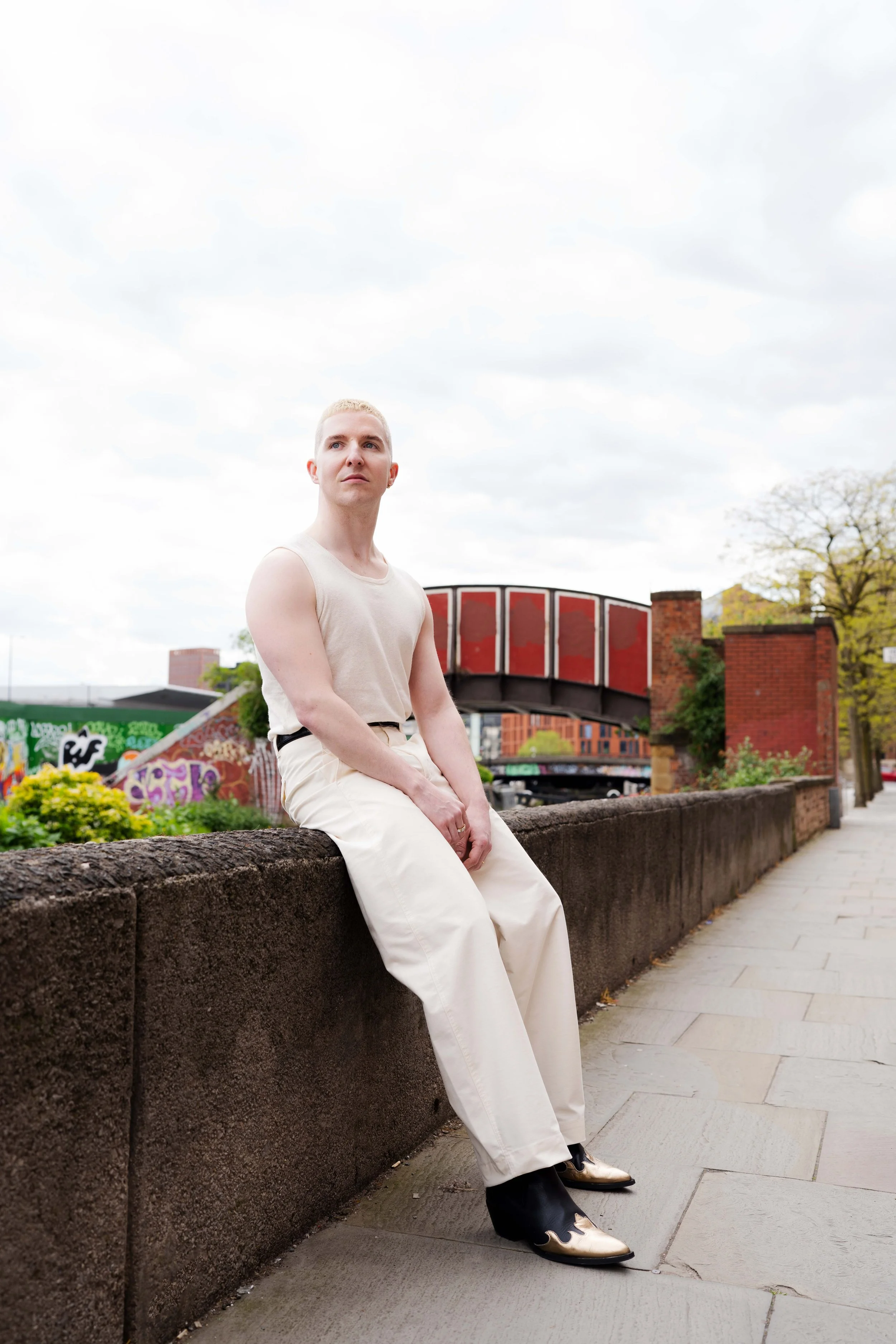 A person with short hair and light skin is sitting on a stone wall along a sidewalk, looking into the distance. The person is wearing a sleeveless top, wide-leg pants, and cowboy boots with a metallic finish. In the background, there is a bridge with