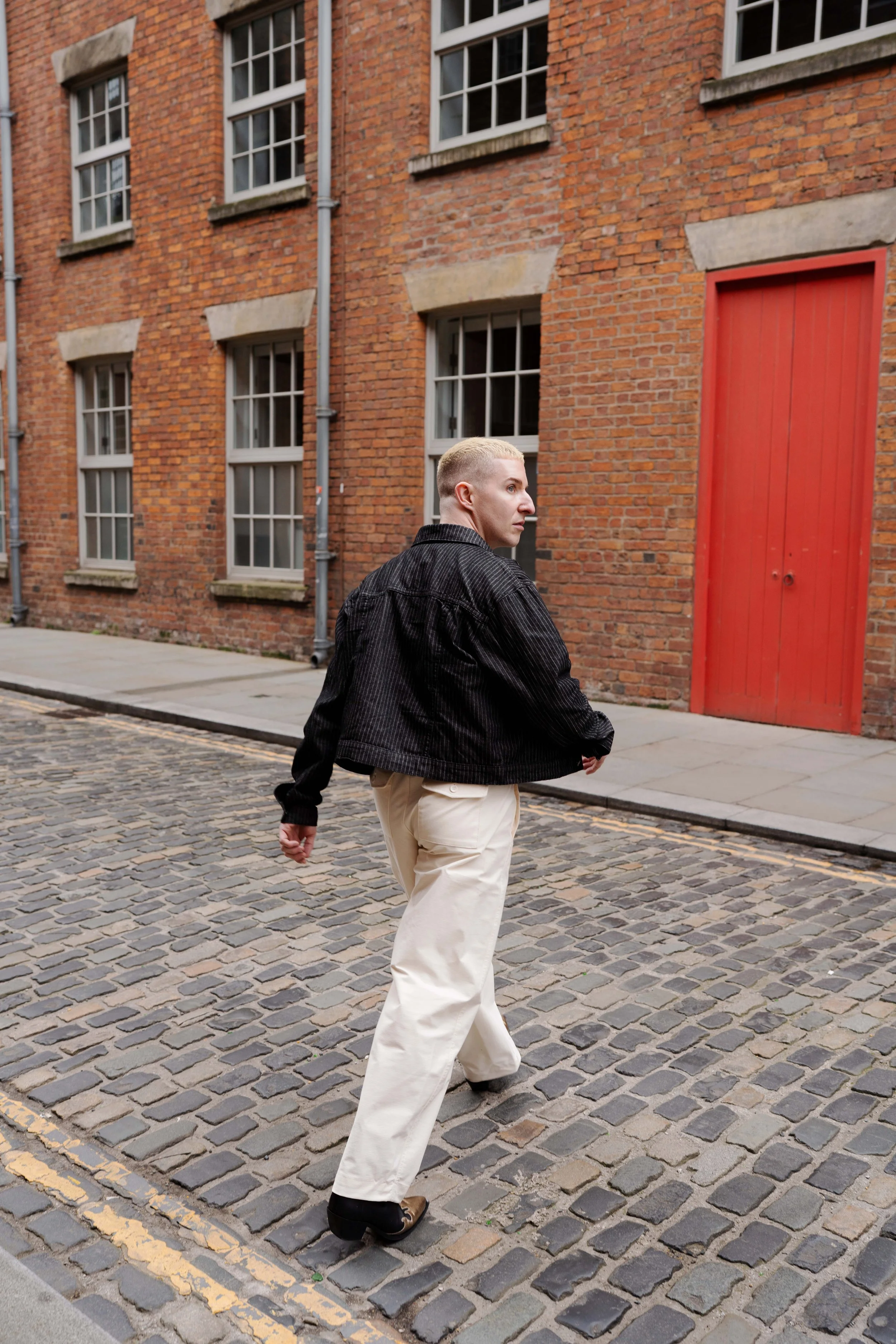 A person with blonde hair wearing a black jacket and cream-colored pants walking on a cobblestone street beside a brick building with multiple windows and a red door.