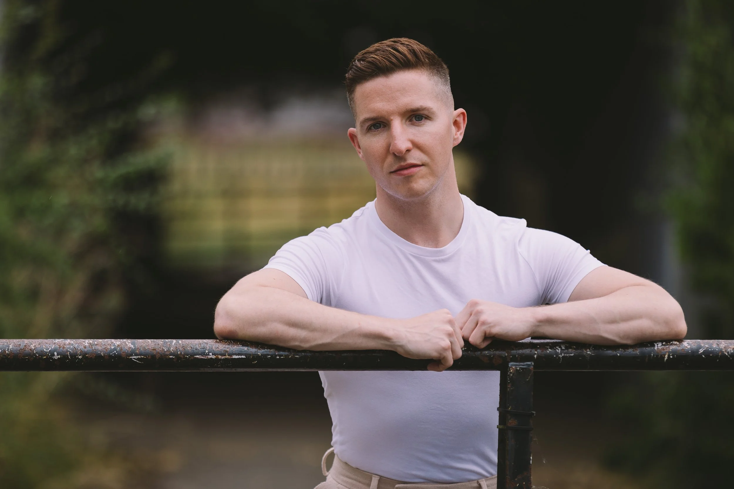 A young man with short brown hair and fair skin leaning on a rusted metal railing outdoors, wearing a white t-shirt, with a blurred green background.