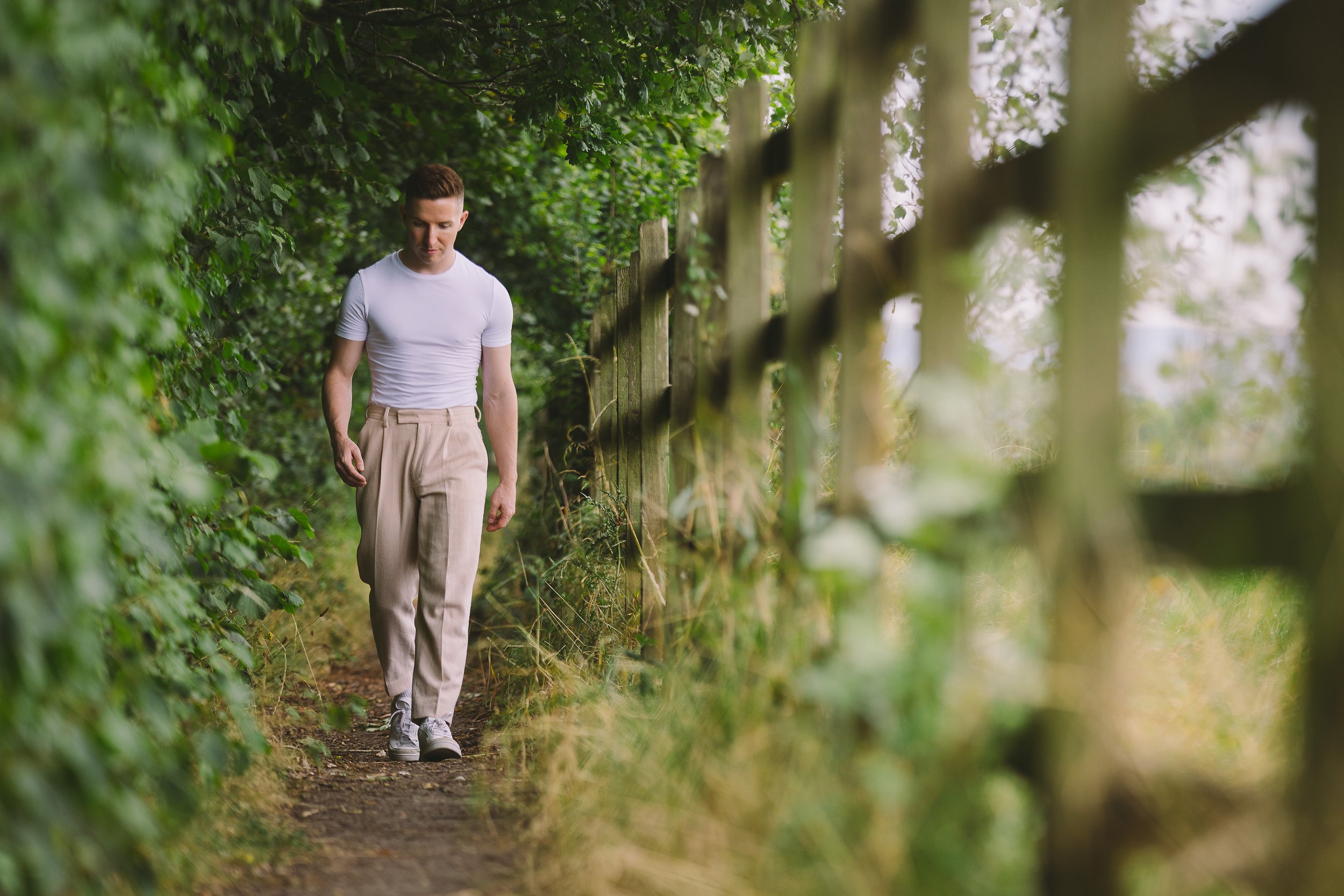 A young man with short hair walking along a narrow dirt path surrounded by green foliage and trees on a cloudy day.