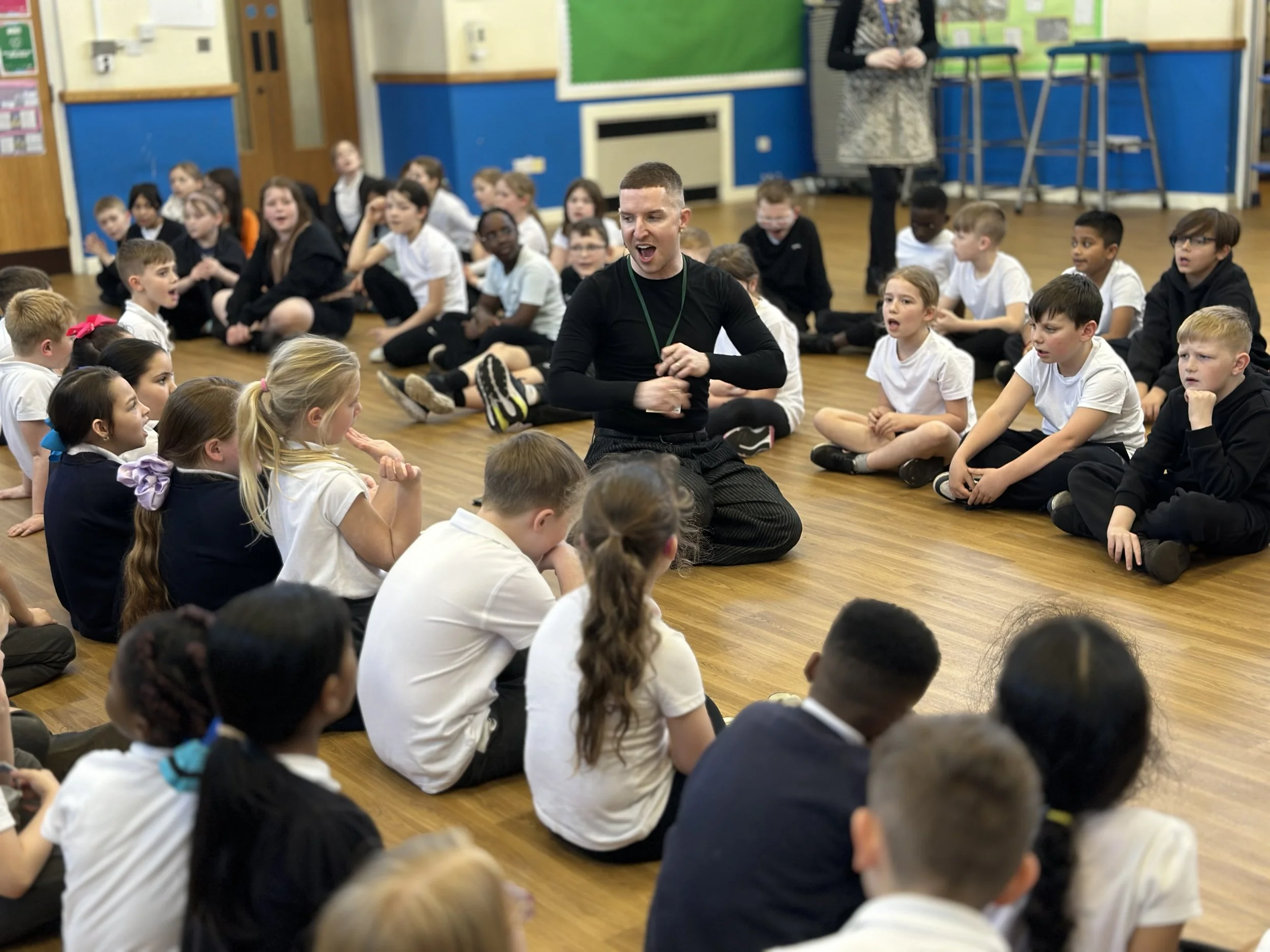 A classroom with children sitting on the floor, listening to a man who is talking to them. The classroom has colorful walls and educational posters.