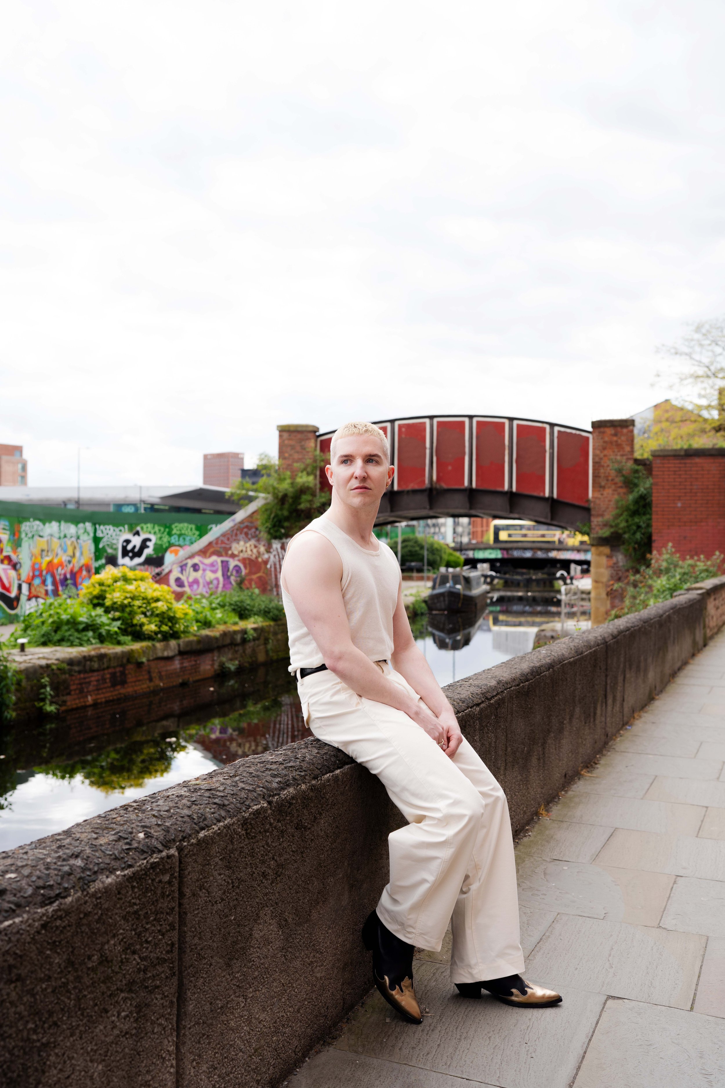 A person sitting on a stone ledge by a canal with colorful graffiti, yellow flowers, and a bridge in the background, wearing a beige sleeveless top and matching pants.