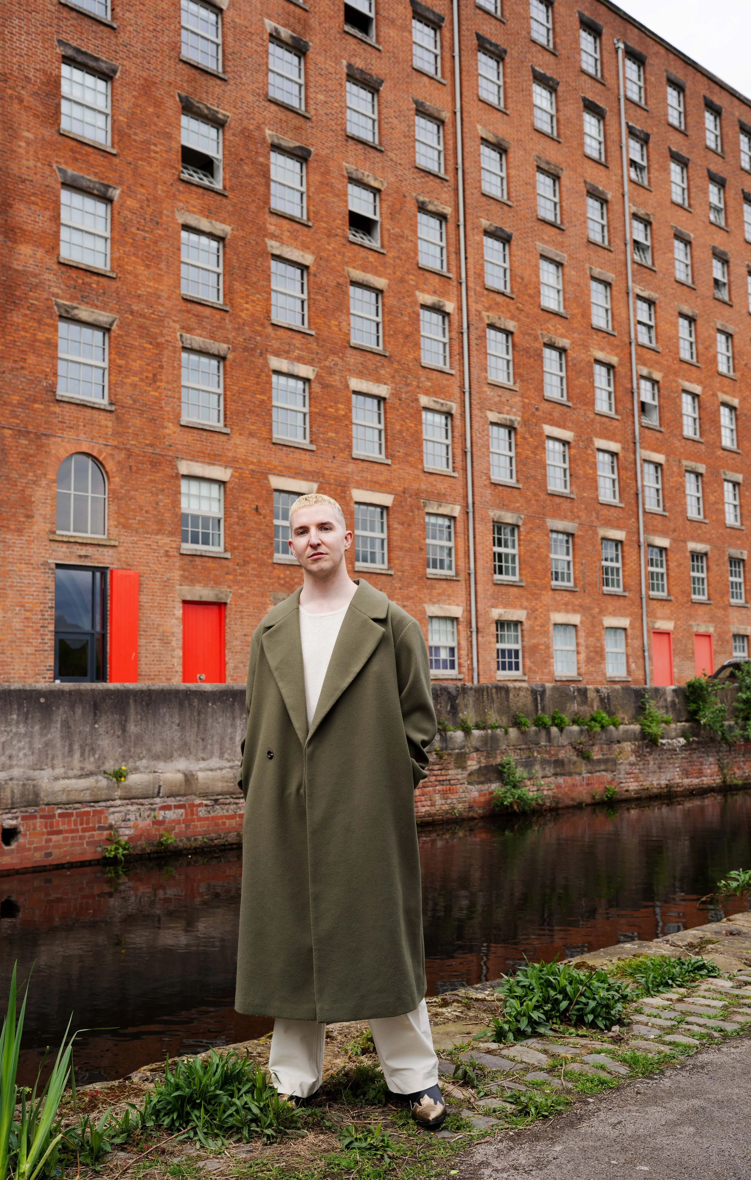 A young man with short blonde hair, wearing a long olive green coat and white pants, standing beside a canal with a brick building in the background.