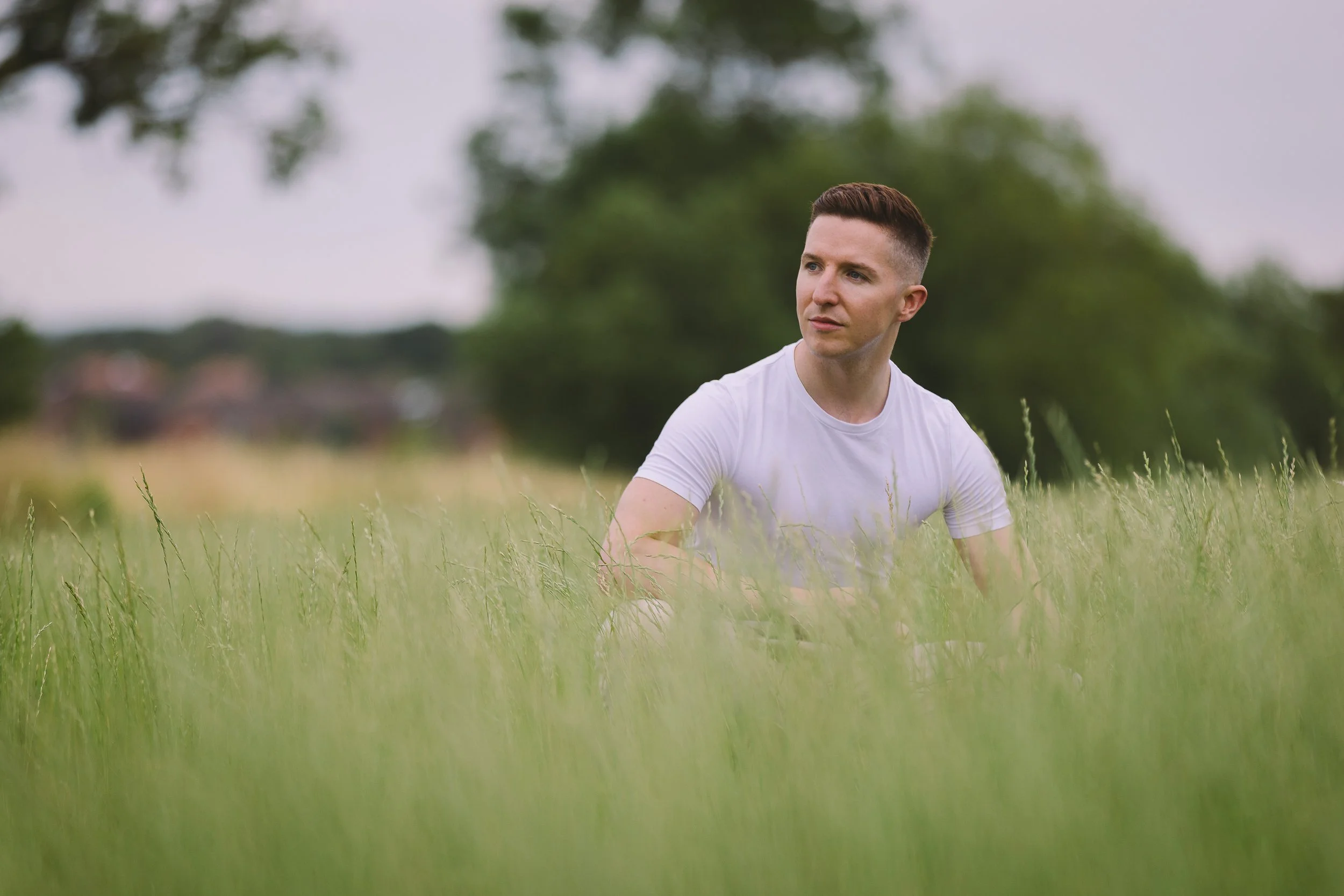 A man with short brown hair and a white t-shirt sitting in a grassy field with trees in the background on a cloudy day.