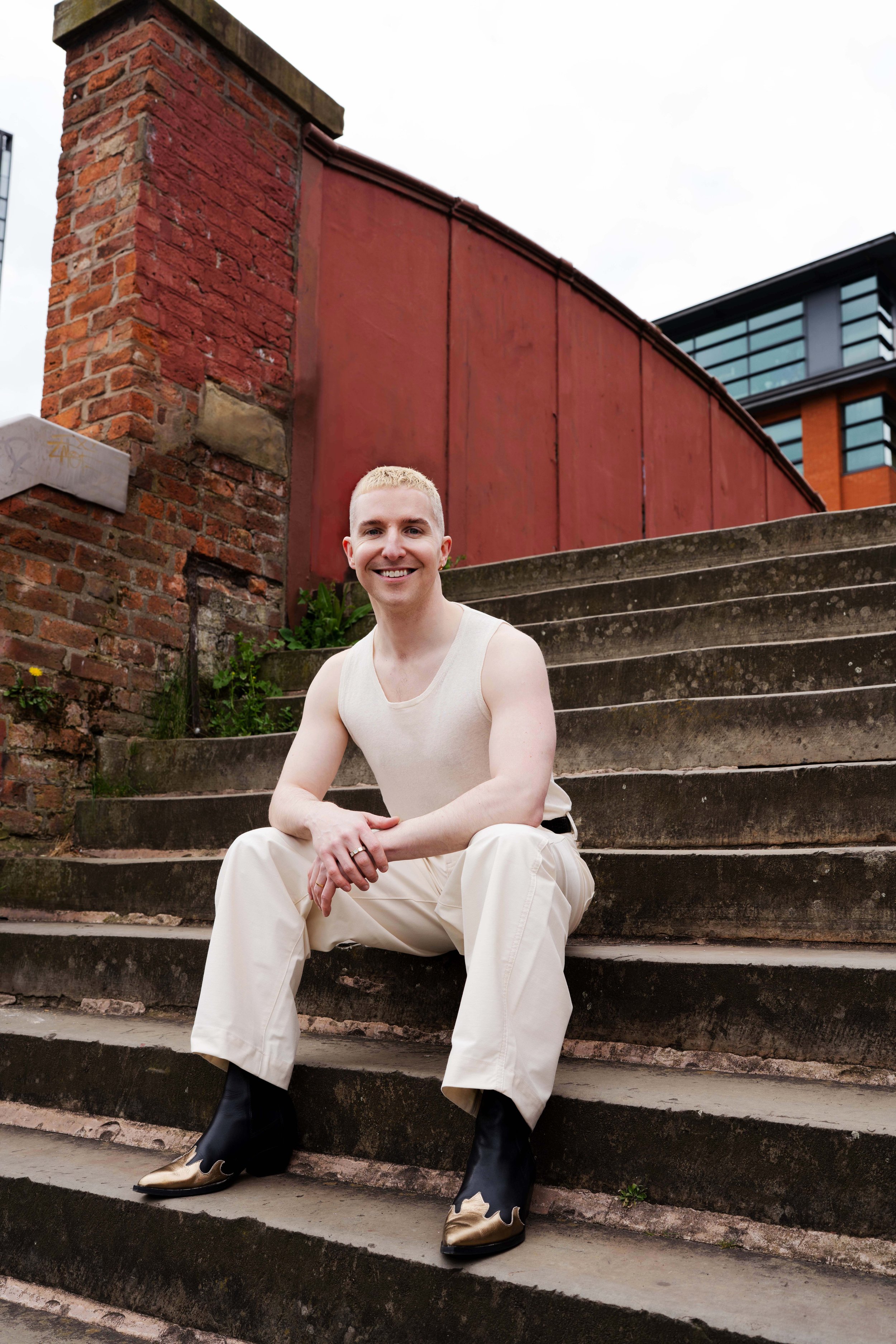 A young man with blond hair, sitting on outdoor concrete stairs, smiling at the camera. He is wearing a white sleeveless shirt, cream-colored pants, and black and gold cowboy boots. Behind him are red brick buildings and a larger modern building with