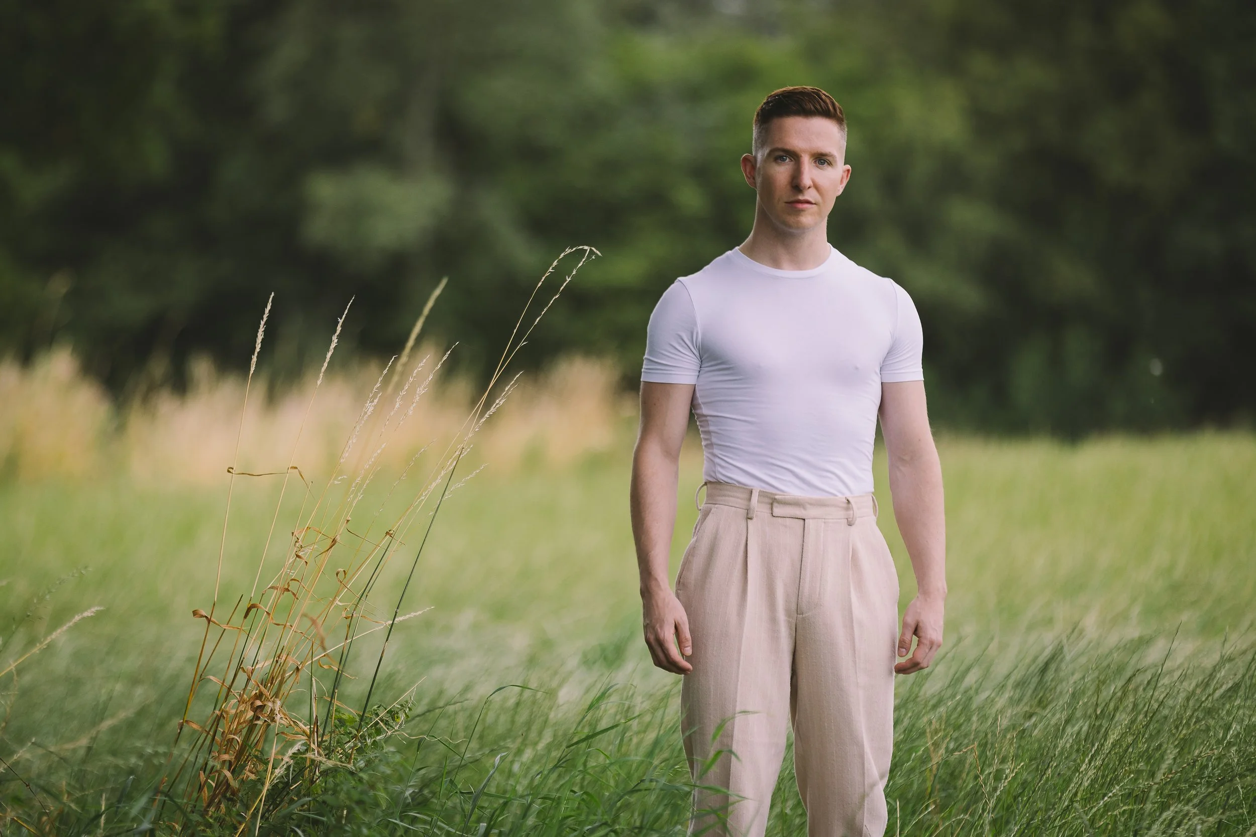 A young man with short brown hair and fair skin stands in a grassy field with blurred green trees in the background. He is wearing a fitted white t-shirt and beige high-waisted pants, and looks directly at the camera with a neutral expression.