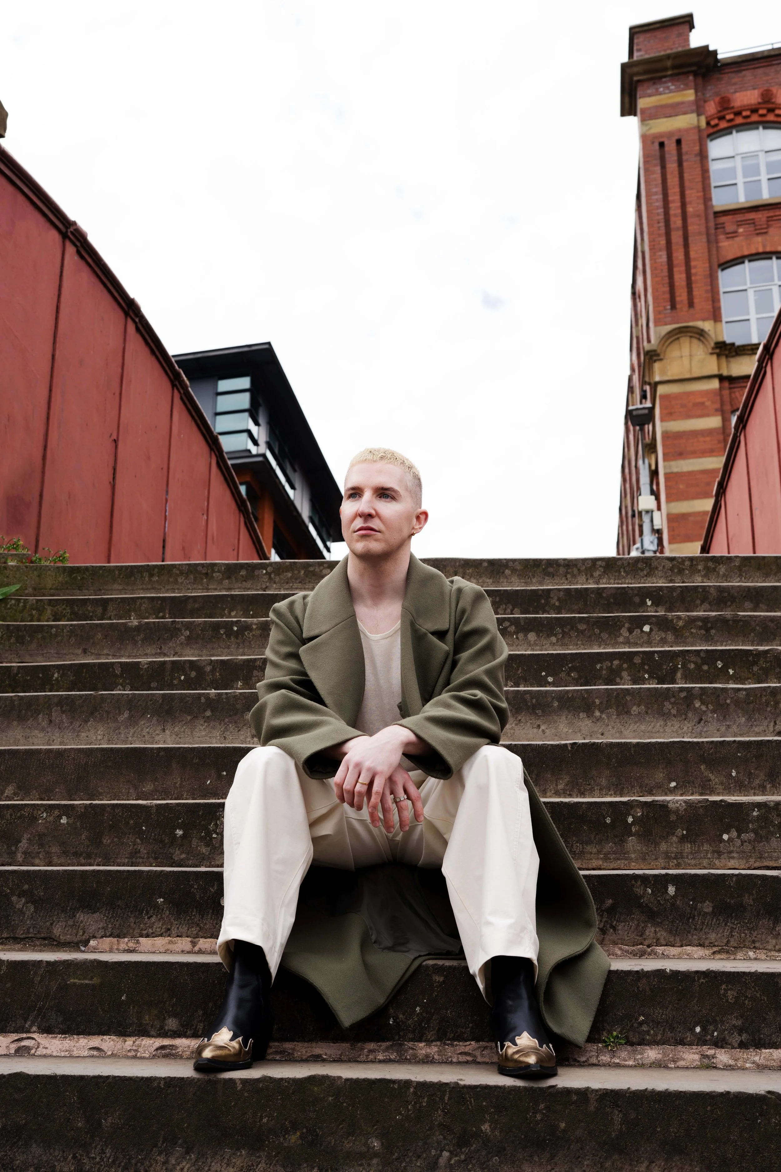A young man with blonde hair sitting on outdoor concrete stairs, wearing a long green coat, cream-colored pants, and black and gold sneakers, with a background of brick and modern buildings.