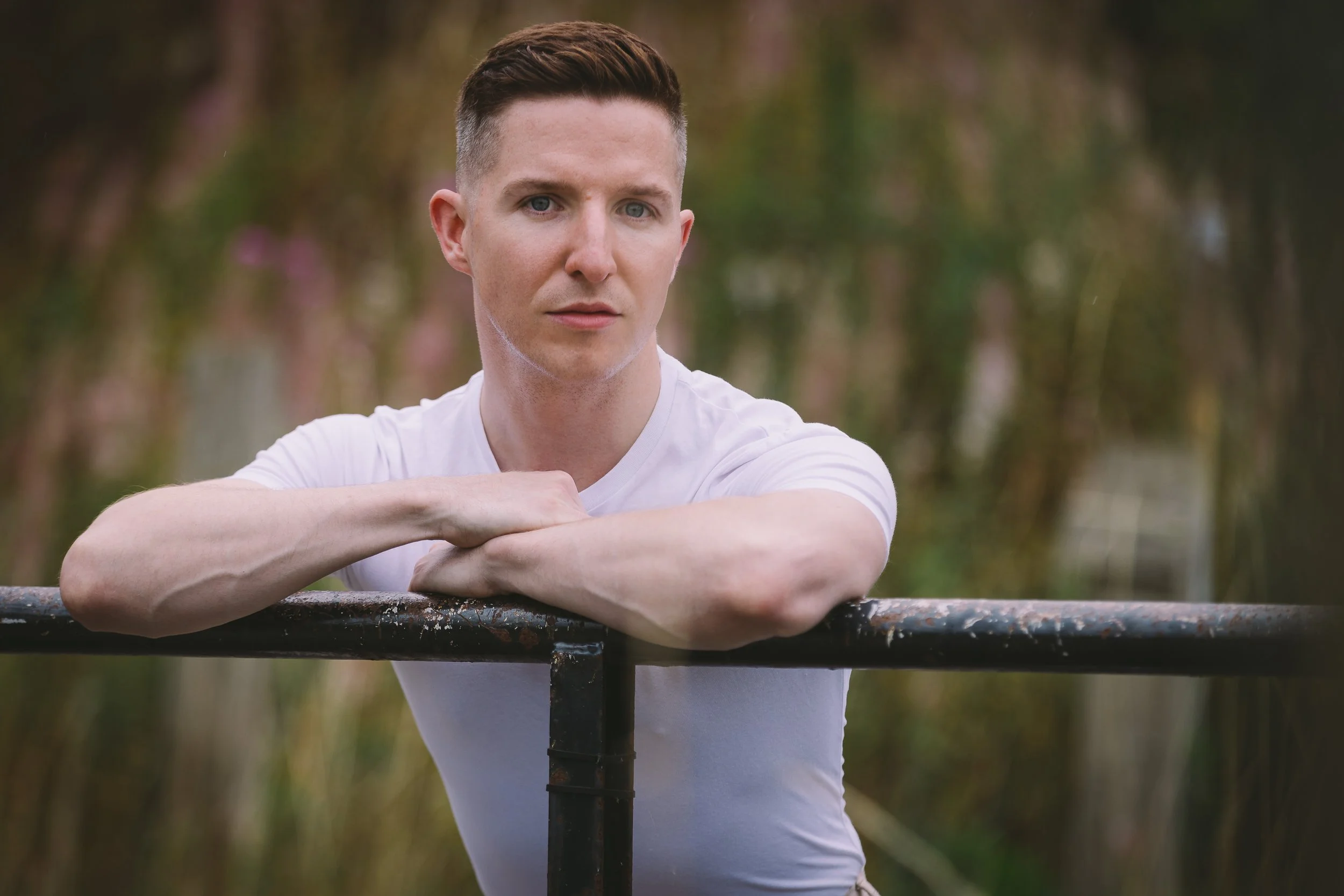 A young man with short brown hair and blue eyes resting his arms on a metal railing outdoors, with a blurred green and pink background.