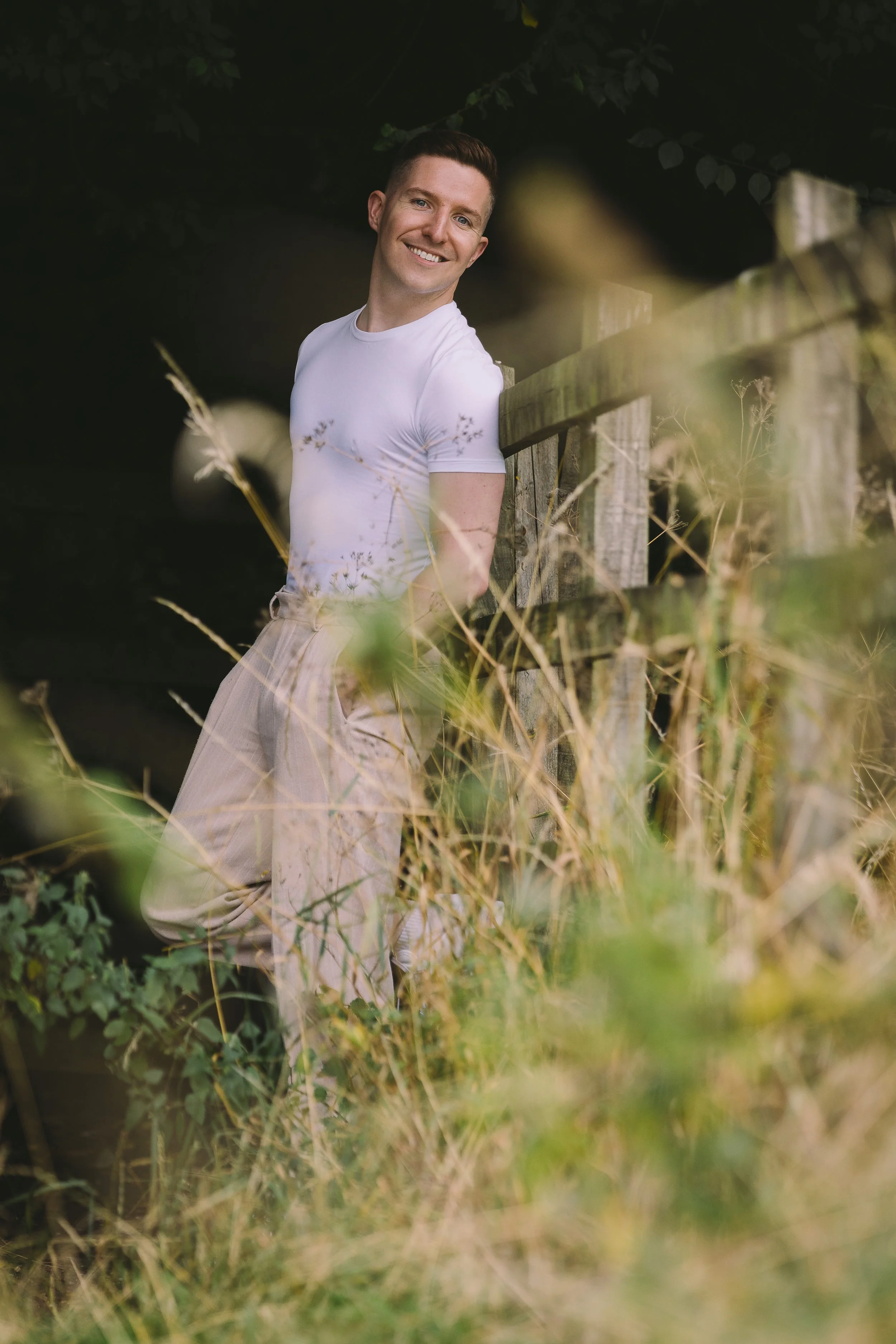 A smiling man in a white t-shirt standing outdoors, leaning on a wooden fence, surrounded by tall grass and greenery.