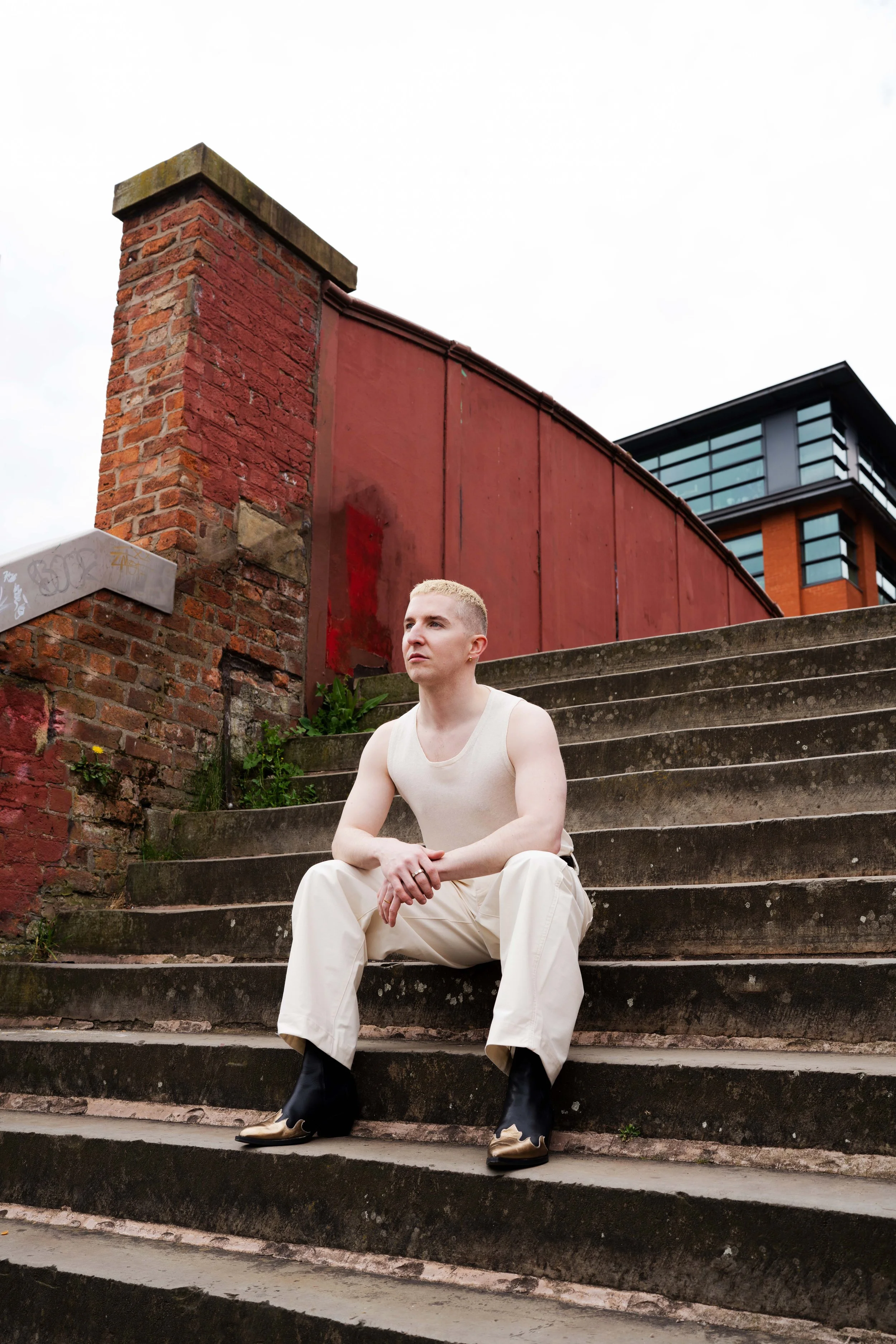 A young person with short blonde hair and a serious expression, sitting on concrete stairs outdoors. They are wearing a sleeveless white top, beige pants, and cowboy boots. Behind them, there's a brick chimney, a red wall, and a modern building with 
