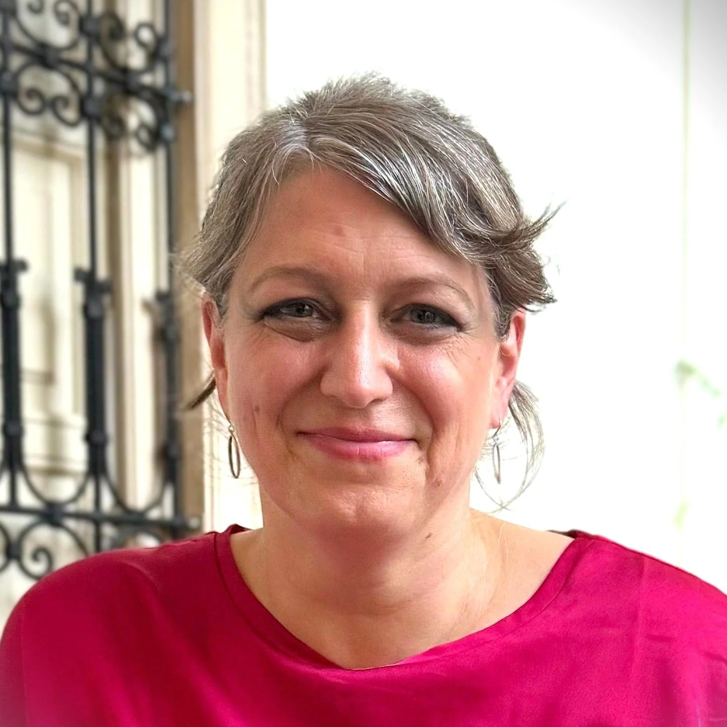 Headshot of Martha Lawton, smiling calmly, in a sunny courtyard in front of a whitewashed wall with a shuttered window behind her