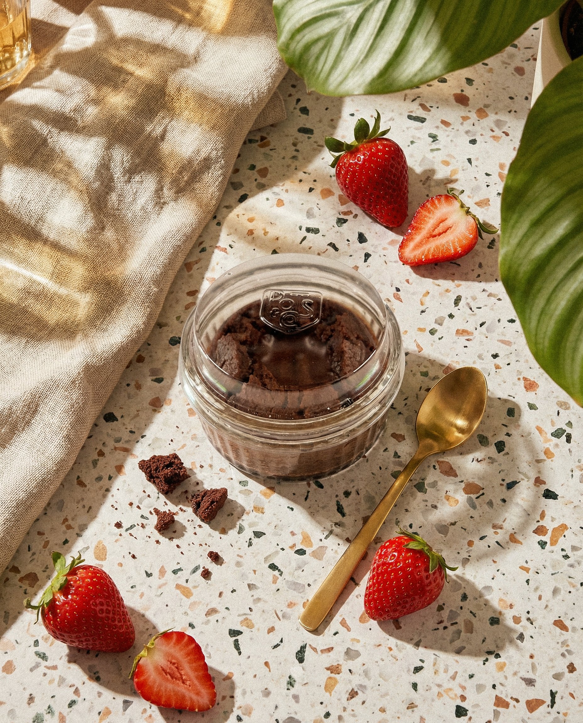 Fresh strawberries, some sliced, on a speckled white tabletop with a jar of chocolate cake in a glass container, a gold spoon, and a beige cloth.