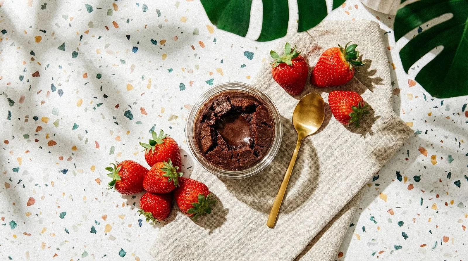A glass jar of chocolate fudge with strawberries and a gold spoon on a beige napkin, on a terrazzo table with green leaves