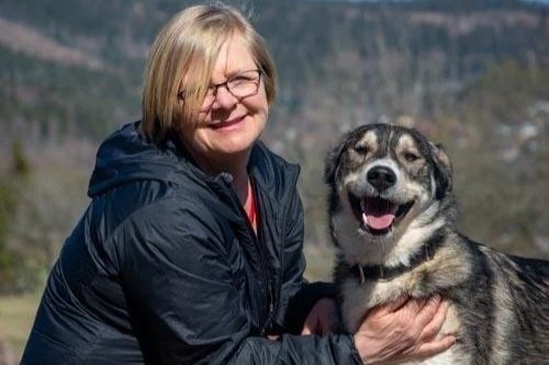 En kvinne i blå jakke holder en grå og svart hund utendørs med fjell i bakgrunnen.