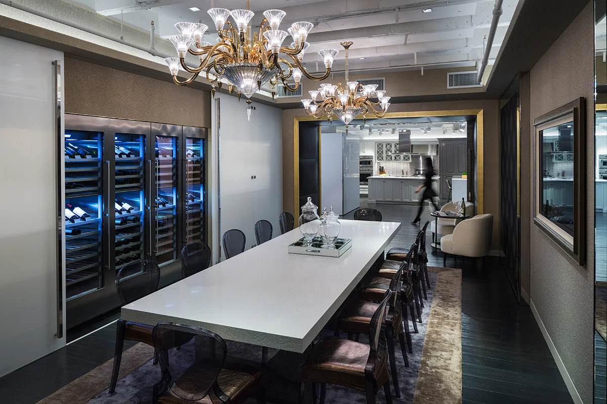 Dining room display with a long white table, wine refrigerators, and chandeliers and a woman walking past. MEP designed by 2L Engineering.
