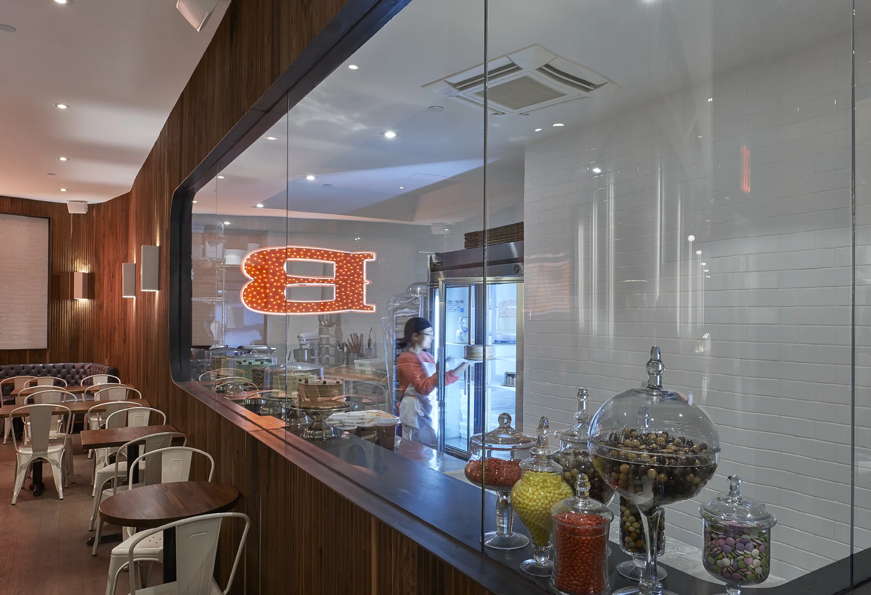 Viewing window to the kitchen of Baked Tribeca where a worker places a cake inside a bright refrigerator; the large B reflected on the glass. MEP designed by 2L Engineering.