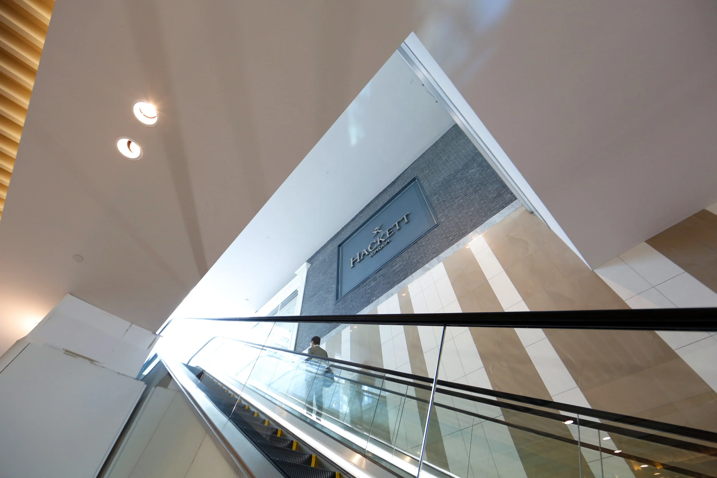 A man standing on an escalator beside the exterior signage of Hackett London in Woodbury Common Premium outlet. MEP designed by 2L Engineering.