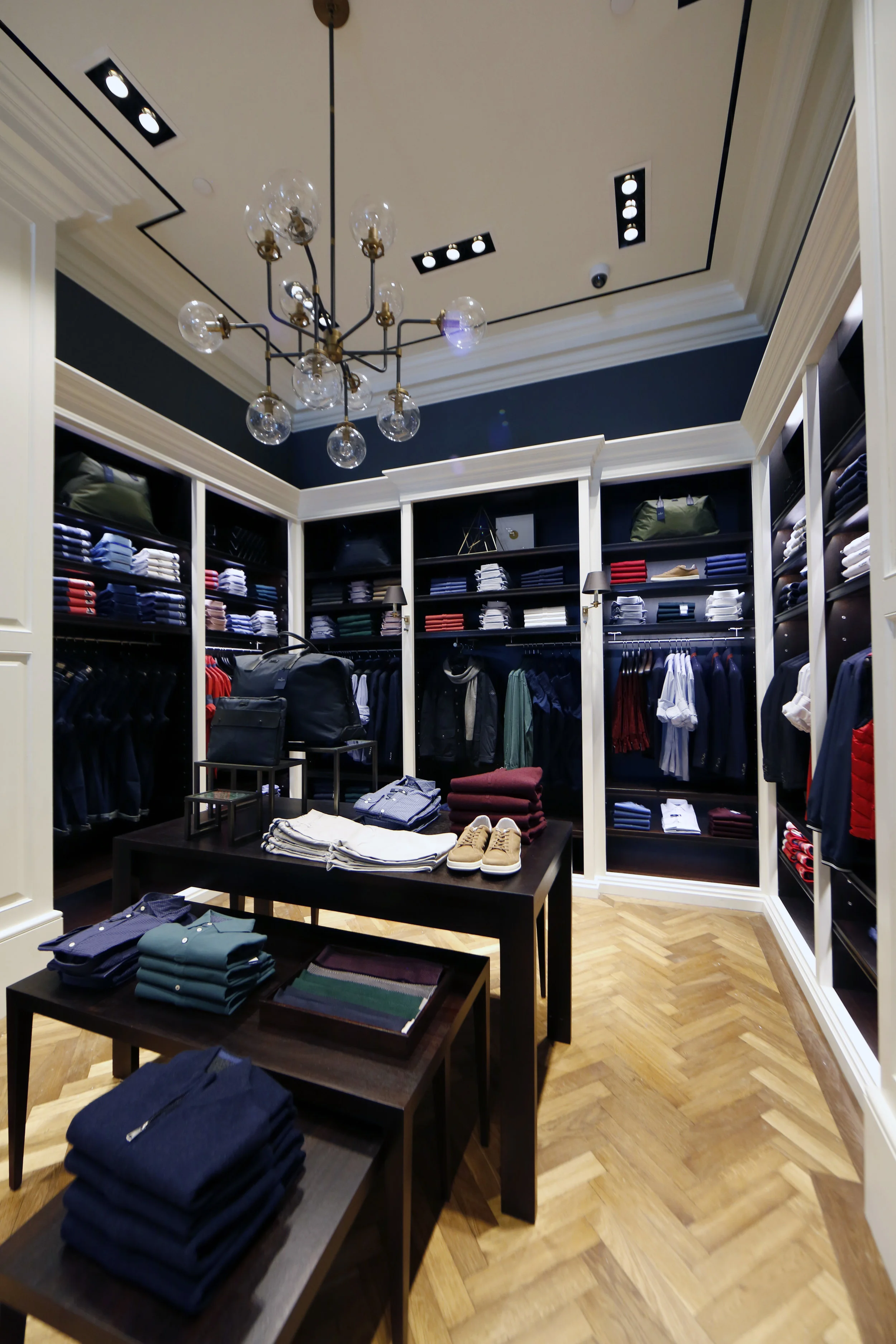 Casual dress shirts and pants on a dark wood display table with tan sneakers and black bags in Hackett London. MEP provided by 2L Engineering.