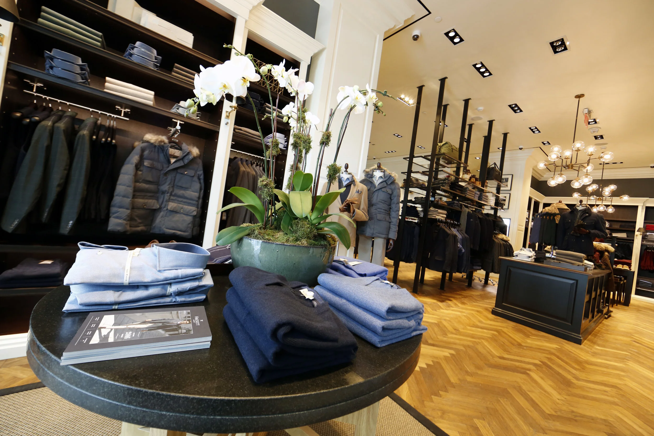 Orchids on a dark wood display table with pullovers, collared shirts, and a Hackett London catalog in Woodbury Common Premium outlet, New York. MEP designed by 2L Engineering.