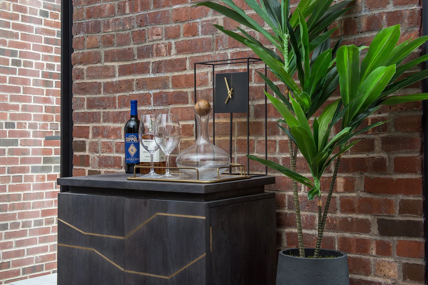 Geometric mini bar with wine, whiskey, glasses, and a minimalistic clock beside a plant against a brick wall. MEP designed by 2L Engineering.