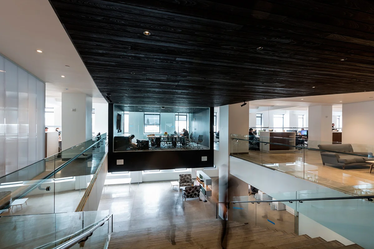 Modern staircase with a brightly lit conference room overhead and dark wood ceiling panels. MEP provided by 2L Engineering.