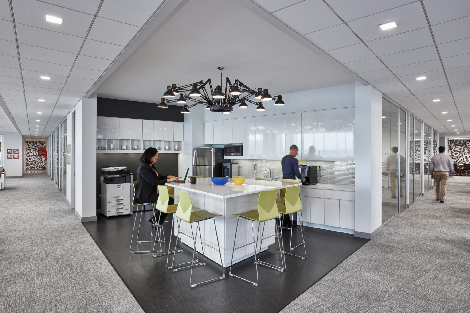 Office kitchenette with a desk lamp chandelier and green bar seats where a woman is working on her laptop as a man makes coffee. MEP provided by 2L Engineering. 