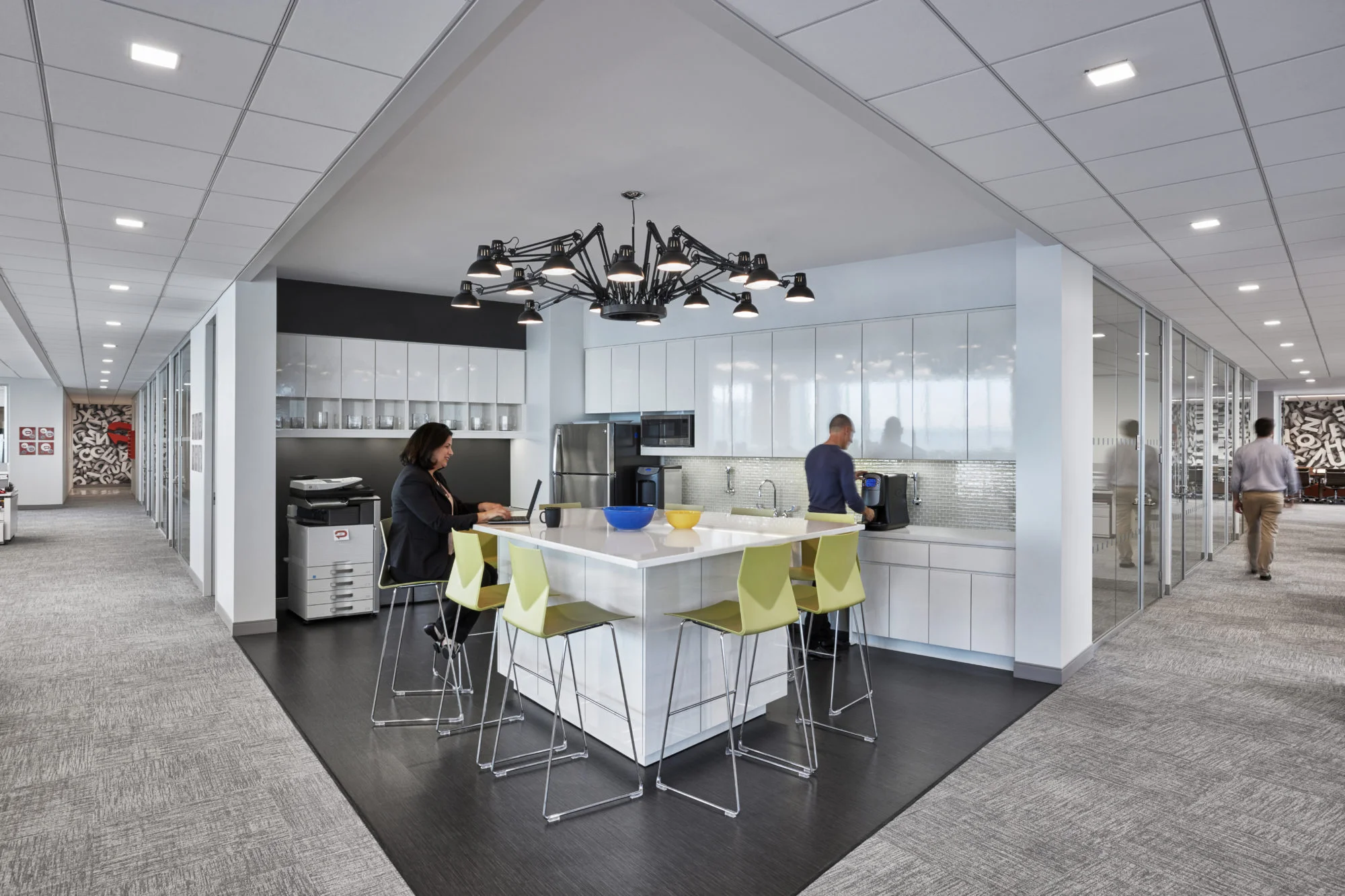 Office kitchenette with a desk lamp chandelier and green bar seats where a woman is working on her laptop as a man makes coffee. MEP provided by 2L Engineering.