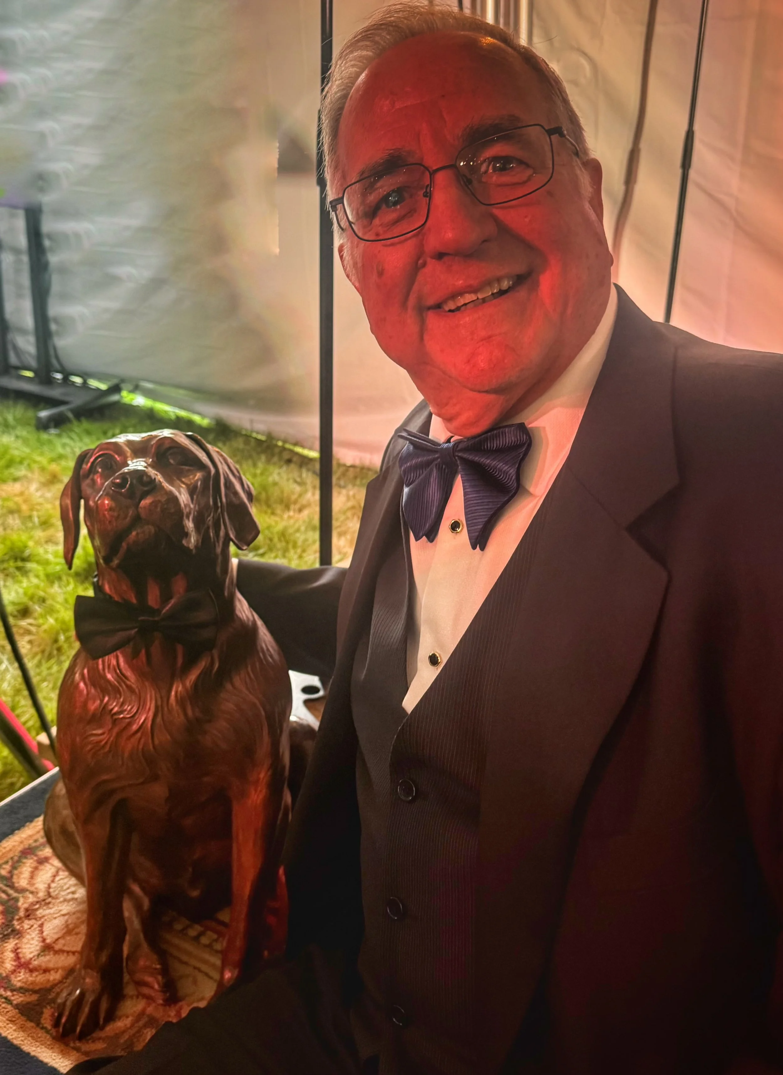 Jake the bronze dog sculpture wearing a bow tie beside the Alpenglow Gala auctioneer in a matching bow tie.