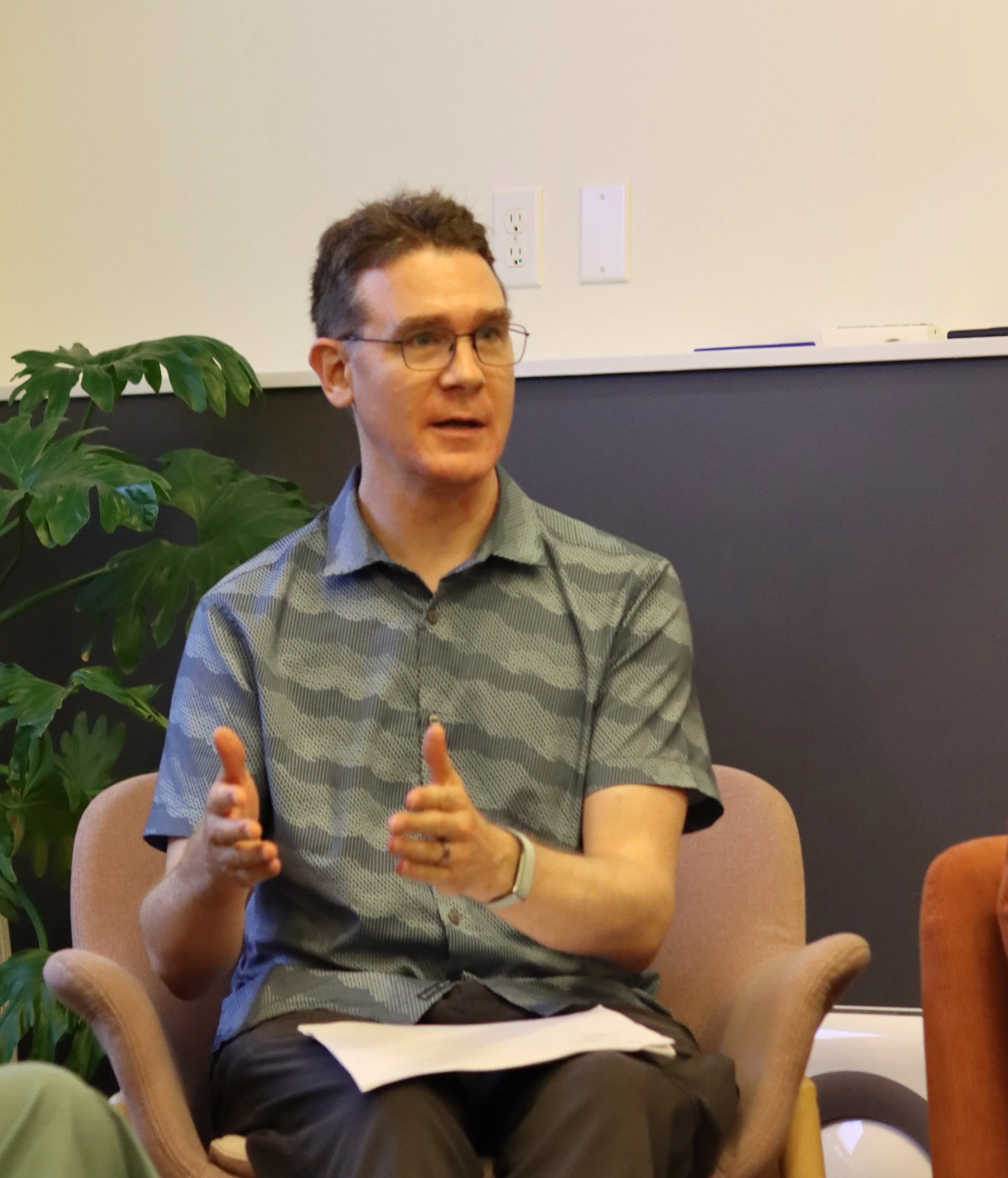 A person wearing a blue shirt and glasses, sitting near a large indoor plant, smiles and gestures while talking.
