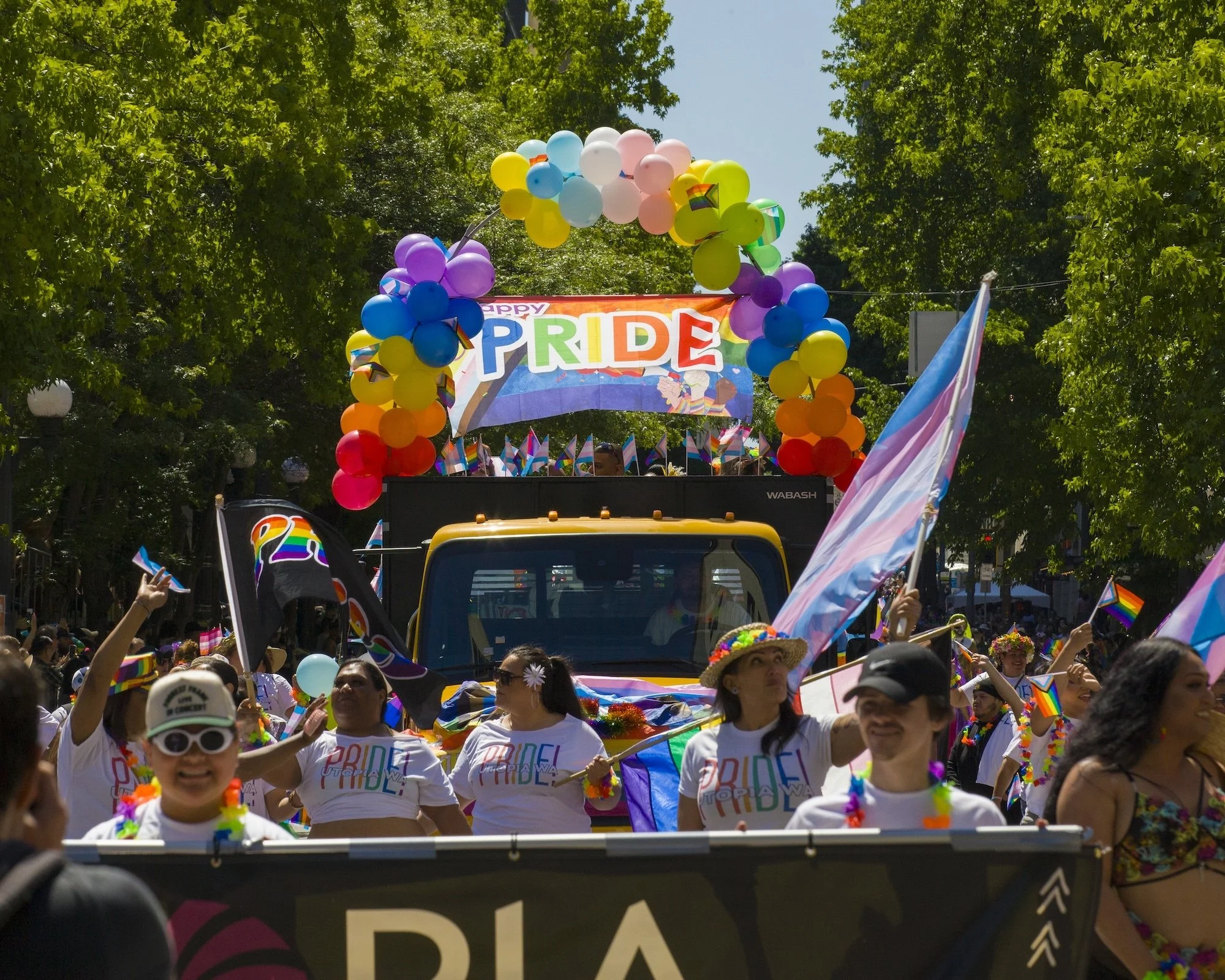People in PRIDE! t-shirts walking in front of a truck on a sunny day. There ae rainbow colored items around the scene.