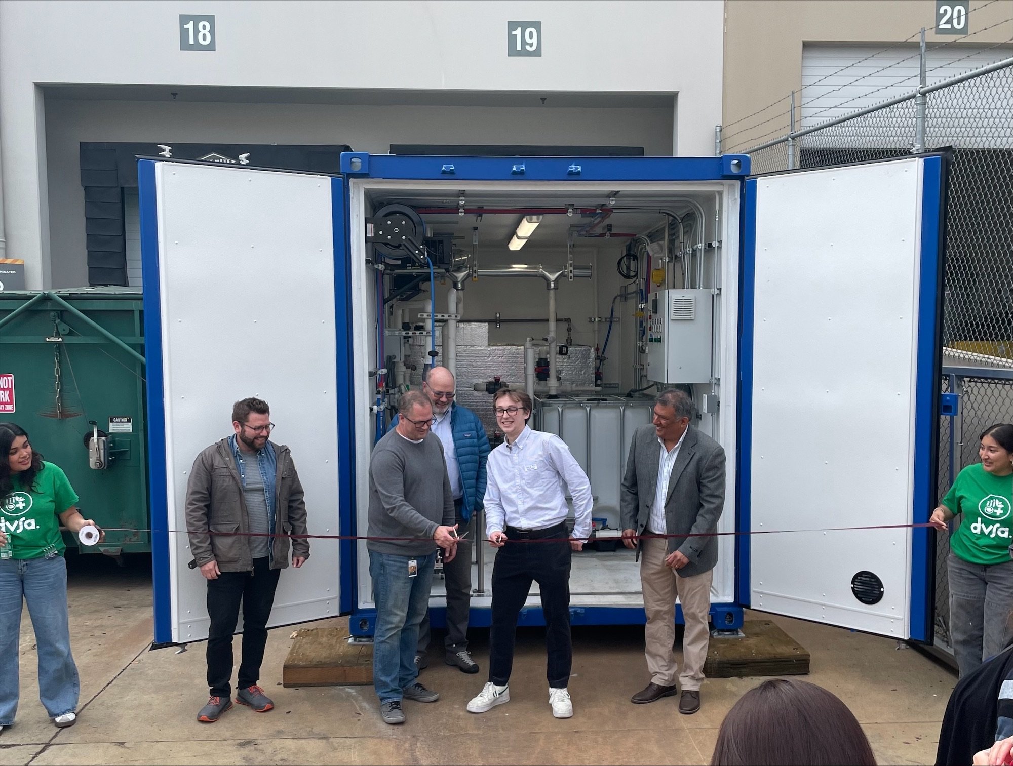 People standing in a row in front of a large outdoor container, holding a ribbon. At the left and right sides, two young people holding the ribbon are wearing green t-shirts that say DVSA.