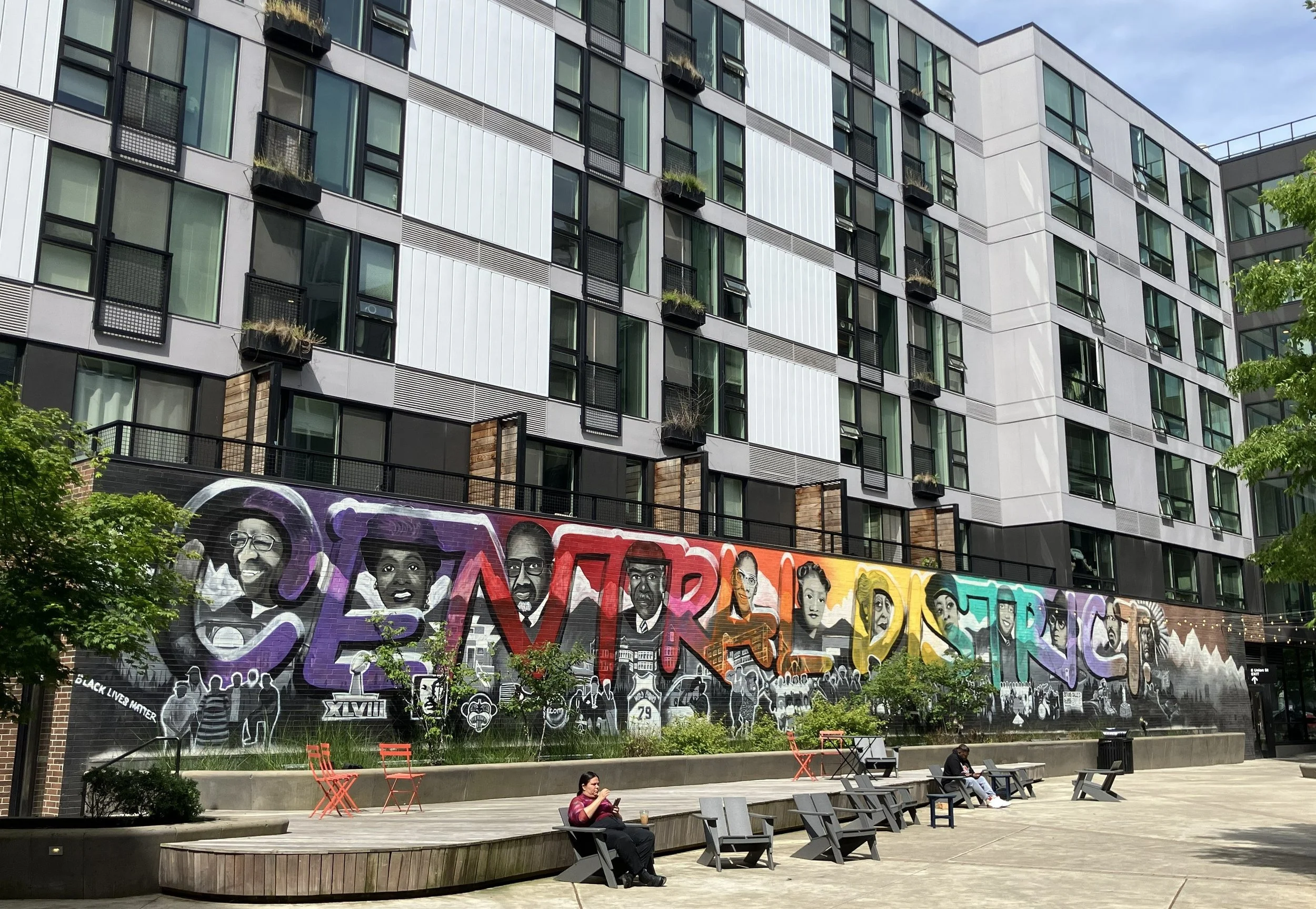 A colorful mural that spells out CENTRAL DISTRICT, with illustrations of Black leaders. Above it is a while apartment building. Infront of it are chairs where people are relaxing.