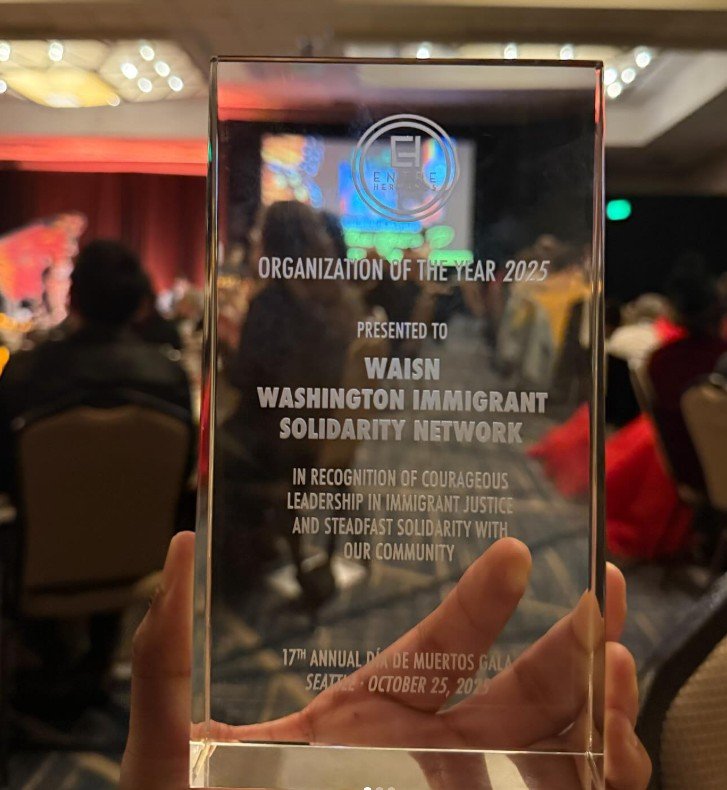 A glass etched award naming WAISN as Organization of the Year, held by a hand, with the banquet in the background.