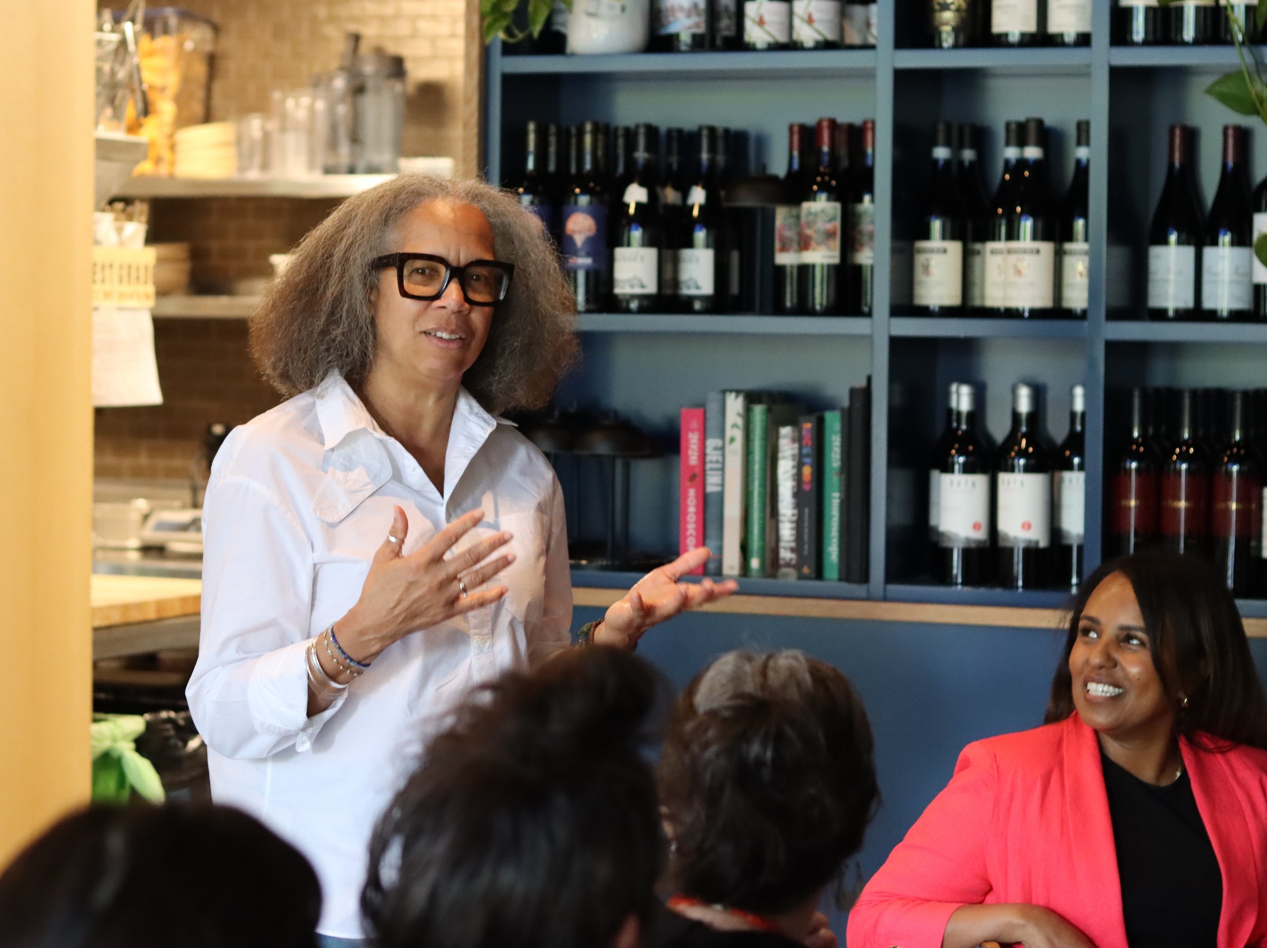 A person wearing a white blouse speaks to a group while a smiling person in a coral-colored jacket listens.