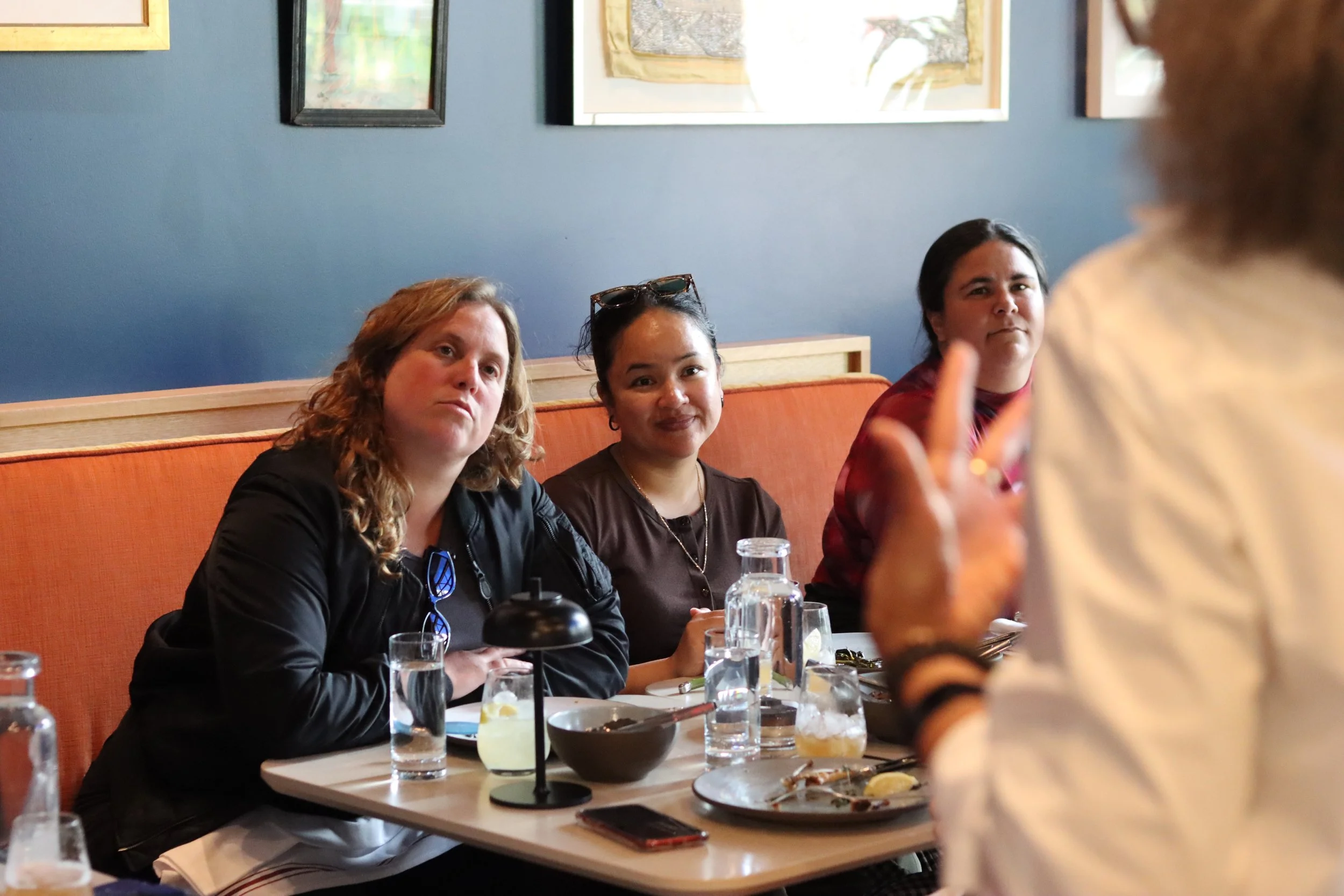 Three people seated at a table as someone in front of them with their back to us gestures.