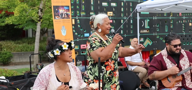 Musicians in Pacific Islander outfits perform as a crowd listens.