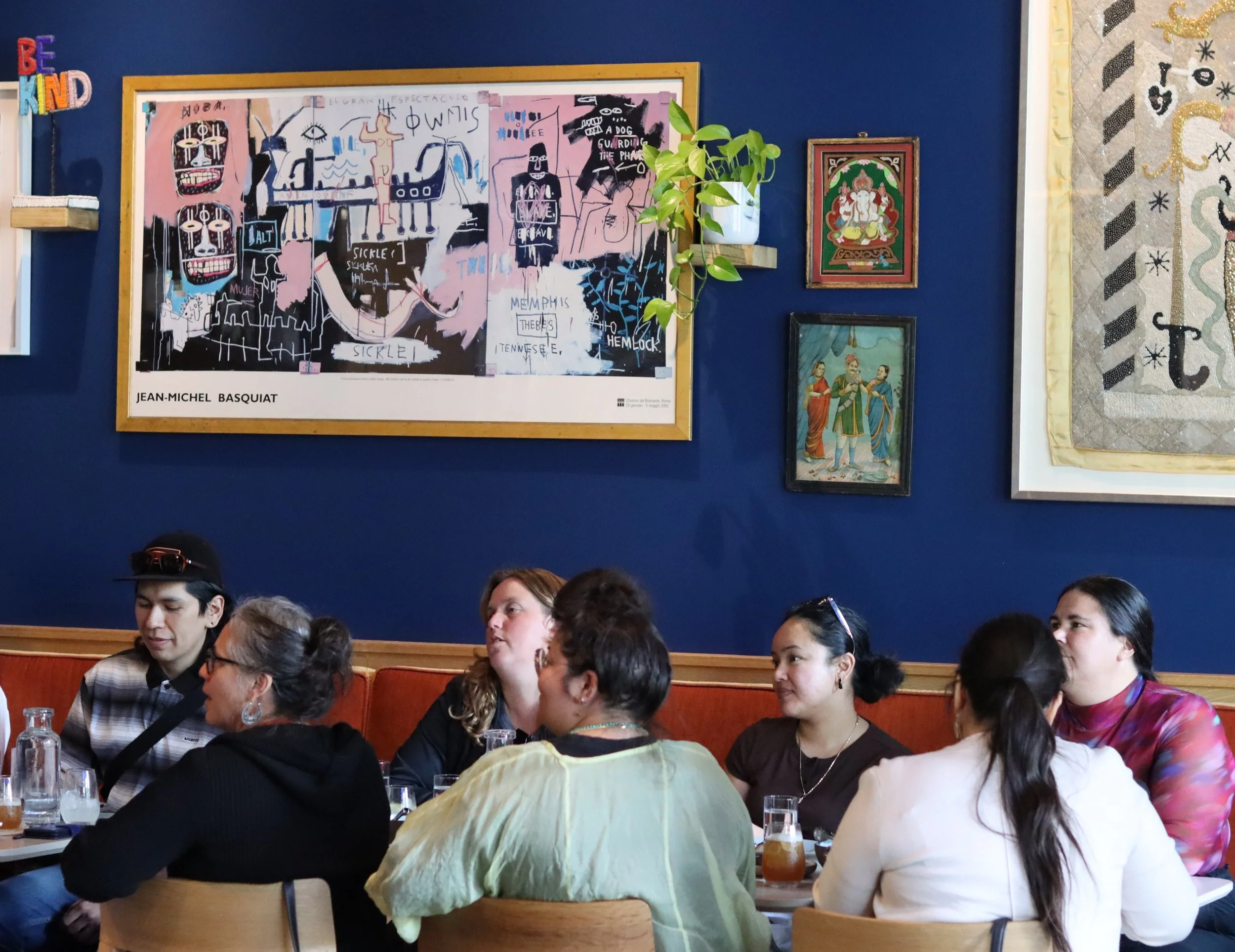 Seven people sit around a table against a blue wall. On the wall are several artworks by Jean-Michel Basquiat and others.