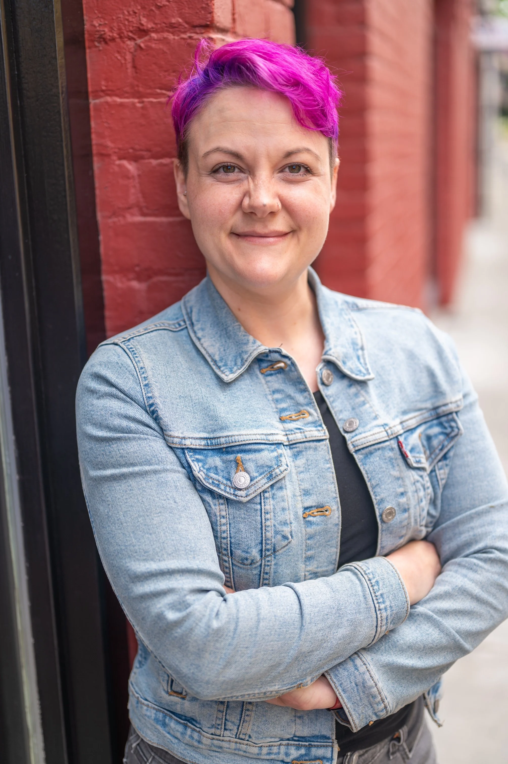 Headshot of a light complexion woman with dyed pink hair wearing a light jean jacket standing in front of a red brick wall