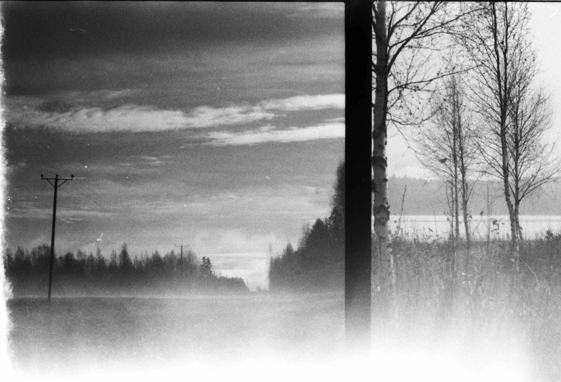 Black and white photograph of a rural landscape with power lines along a dirt road, bare trees in the background, and a cloudy sky.