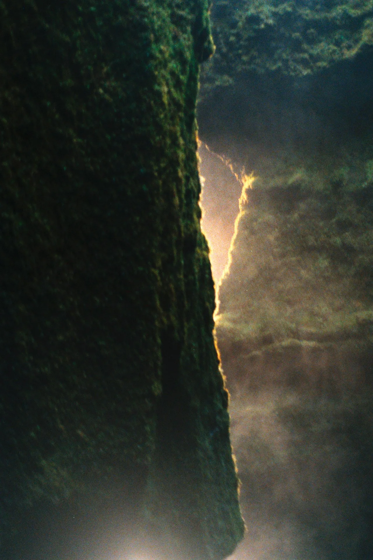 A dark, moss-covered cliff with jagged edges, illuminated by a glowing light behind it, surrounded by misty fog and dramatic cloudy sky.