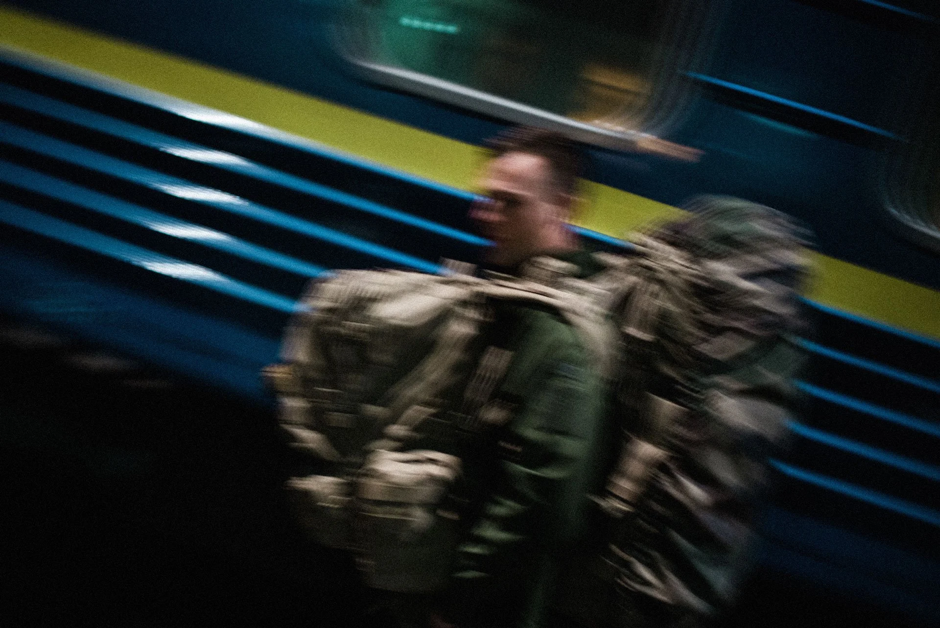 Blurred image of a man in military uniform with a backpack, standing against a blue and green striped background.
