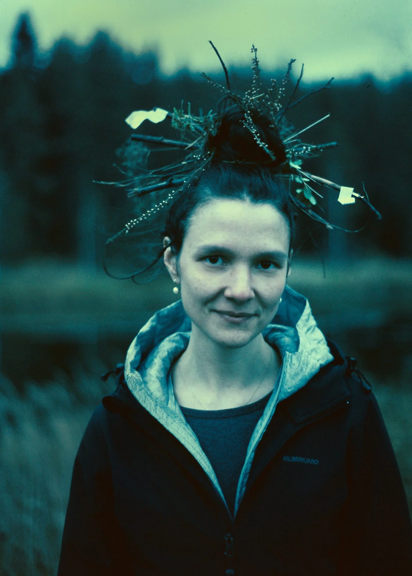 A woman with dark hair in a bun, wearing a jacket with a hood, outdoors in a natural setting during dusk or dawn. She has a decorative headpiece made of sticks, leaves, and small plants.